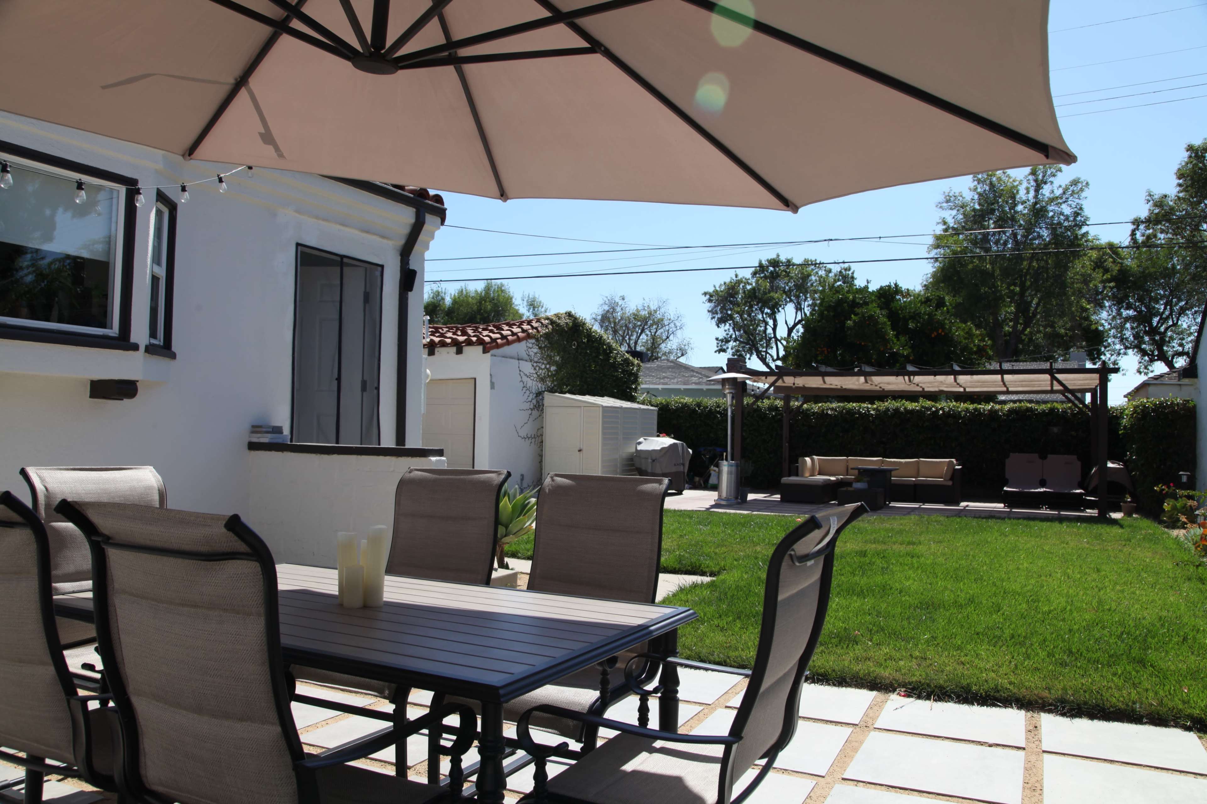 A patio area features a metal dining table with chairs, bordered by a green lawn and a shaded seating area under an umbrella.