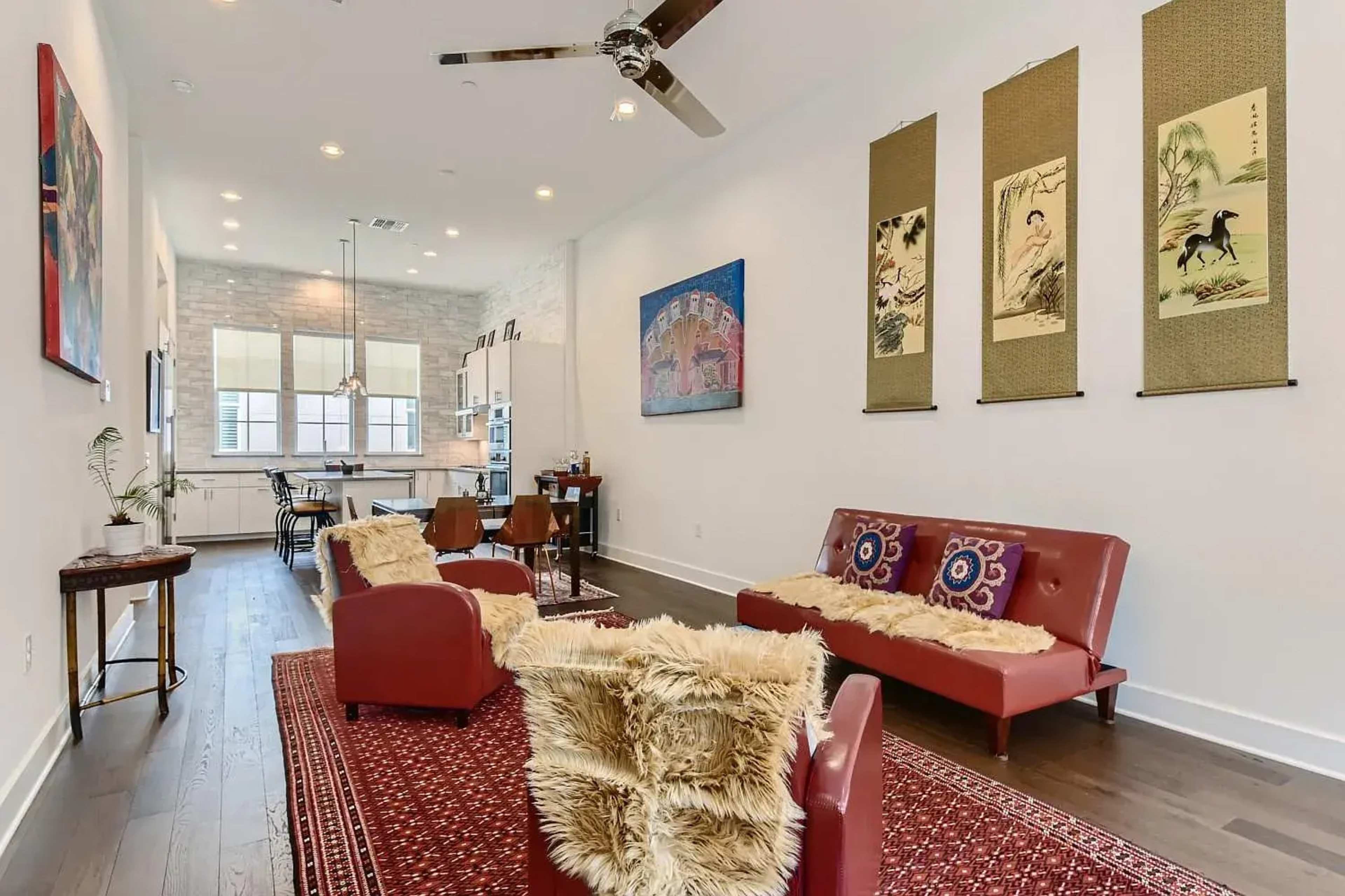 The image shows a living room with red sofas, a patterned rug, and artwork on the walls, leading into a kitchen area visible in the background.