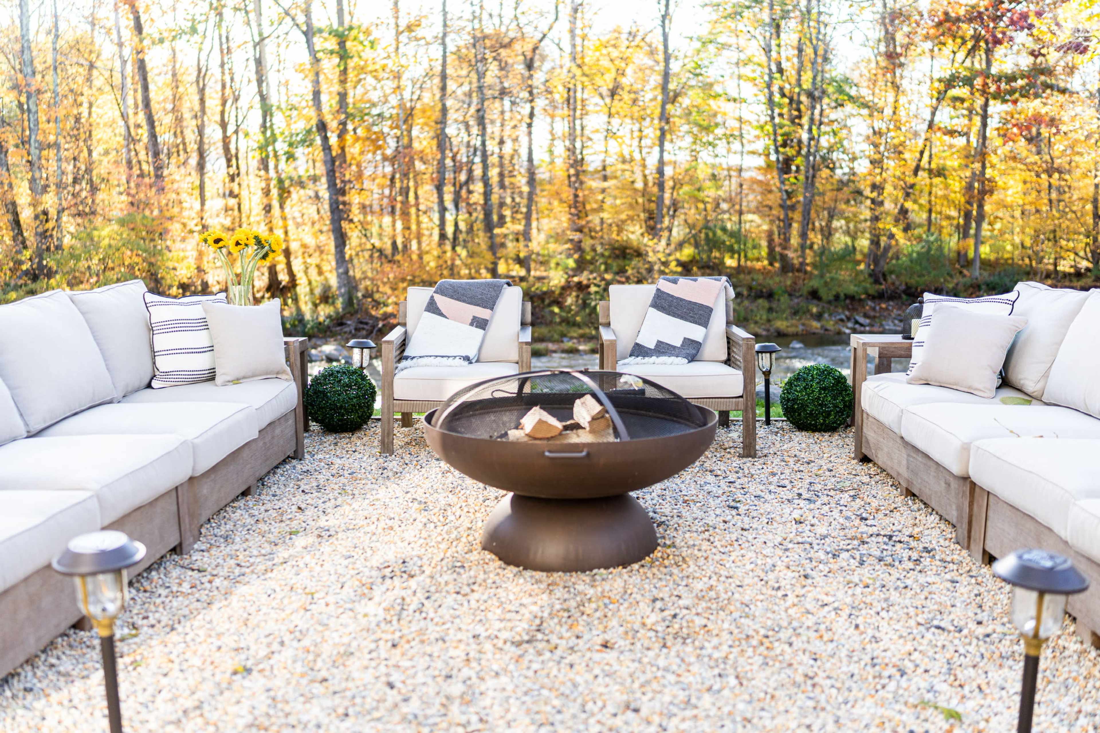 A circular fire pit surrounded by an arrangement of outdoor seating and decorative plants in a gravel area, set against a backdrop of trees with autumn foliage.