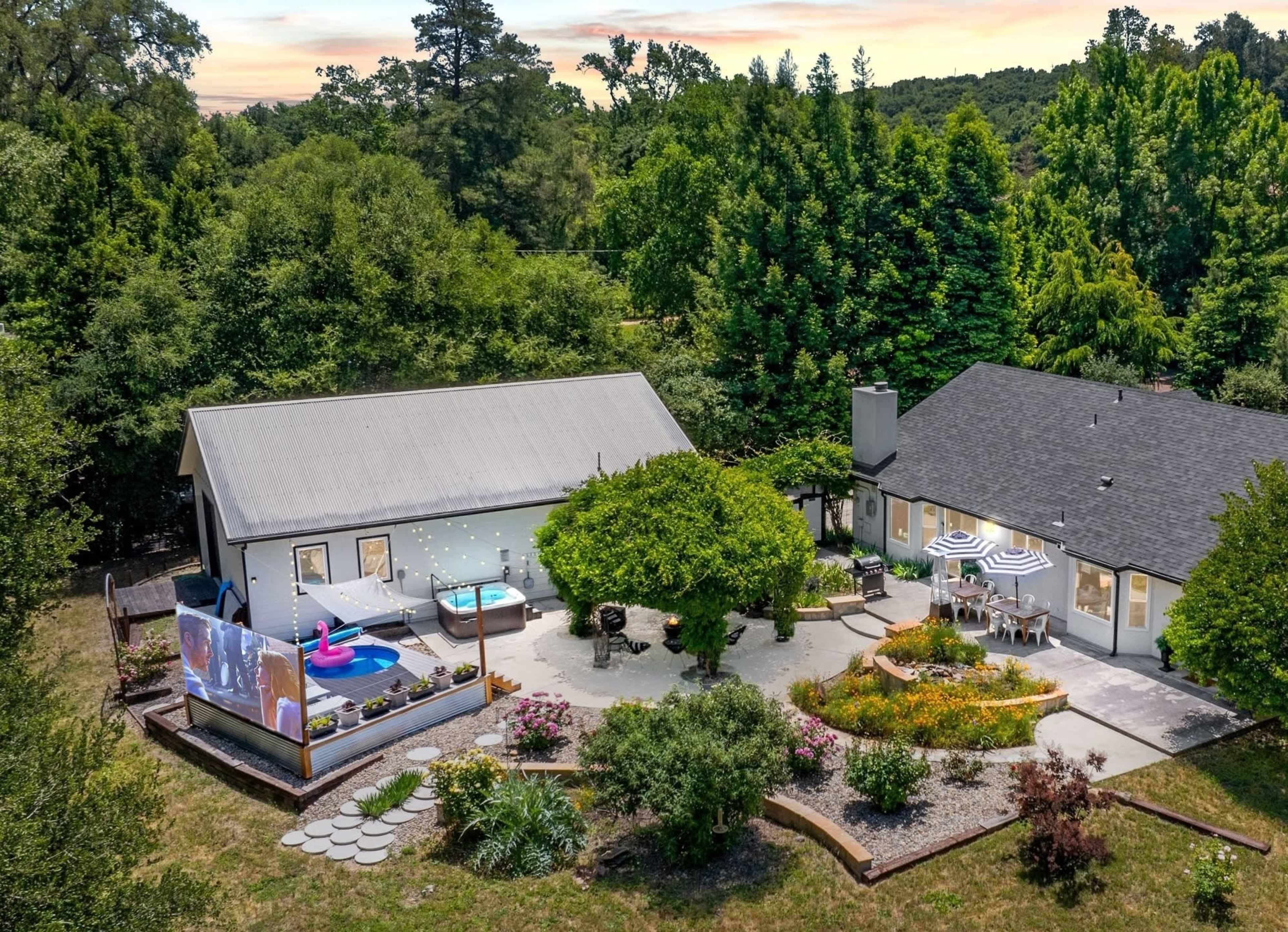 The image shows a spacious backyard with a modern house, a patio area featuring outdoor furniture, a large tree, and a screen displaying a video, surrounded by well-maintained landscaping.