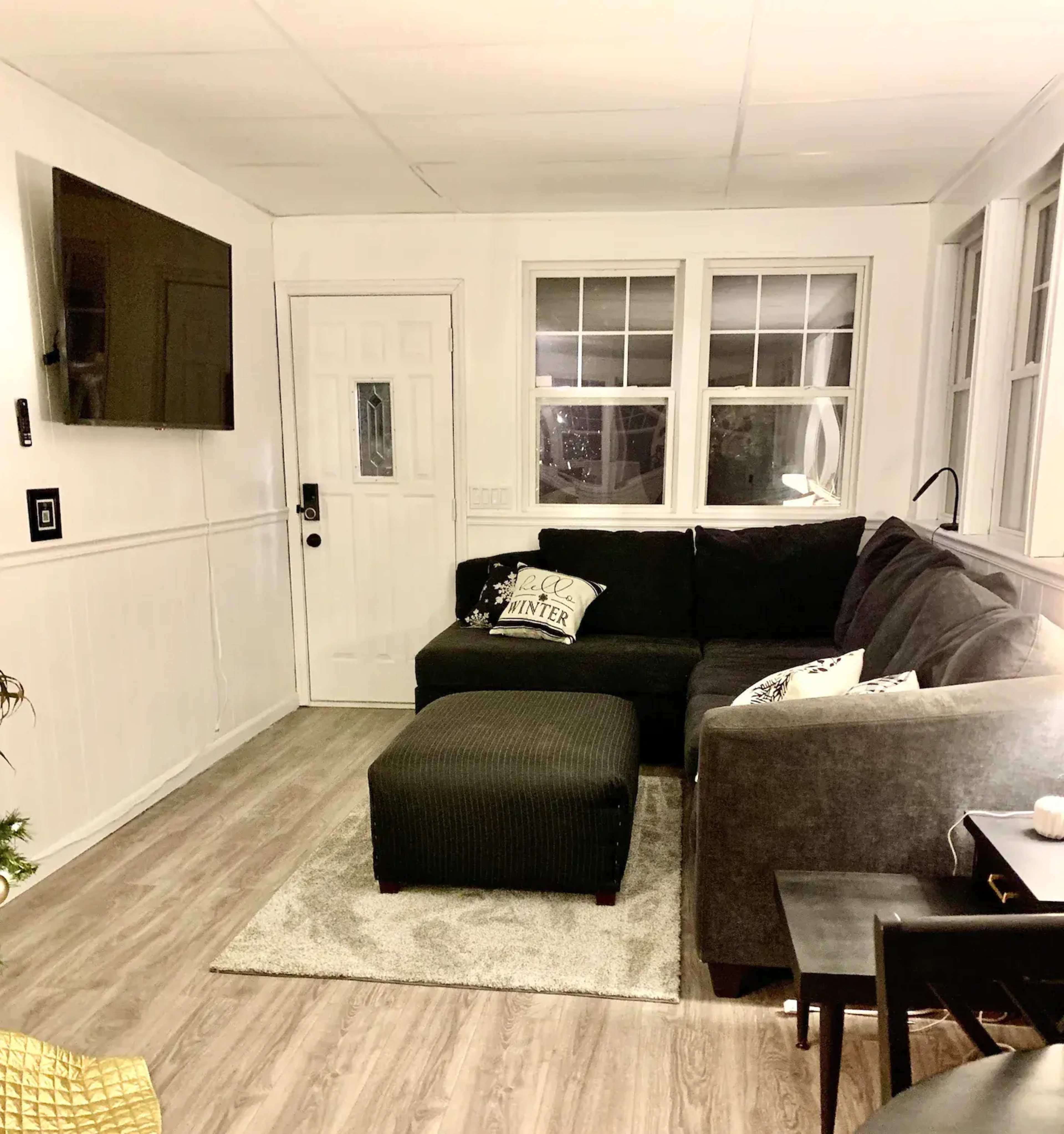 A cozy living room with a dark sectional sofa, a coffee table, and a wall-mounted television near a front door and multiple windows.
