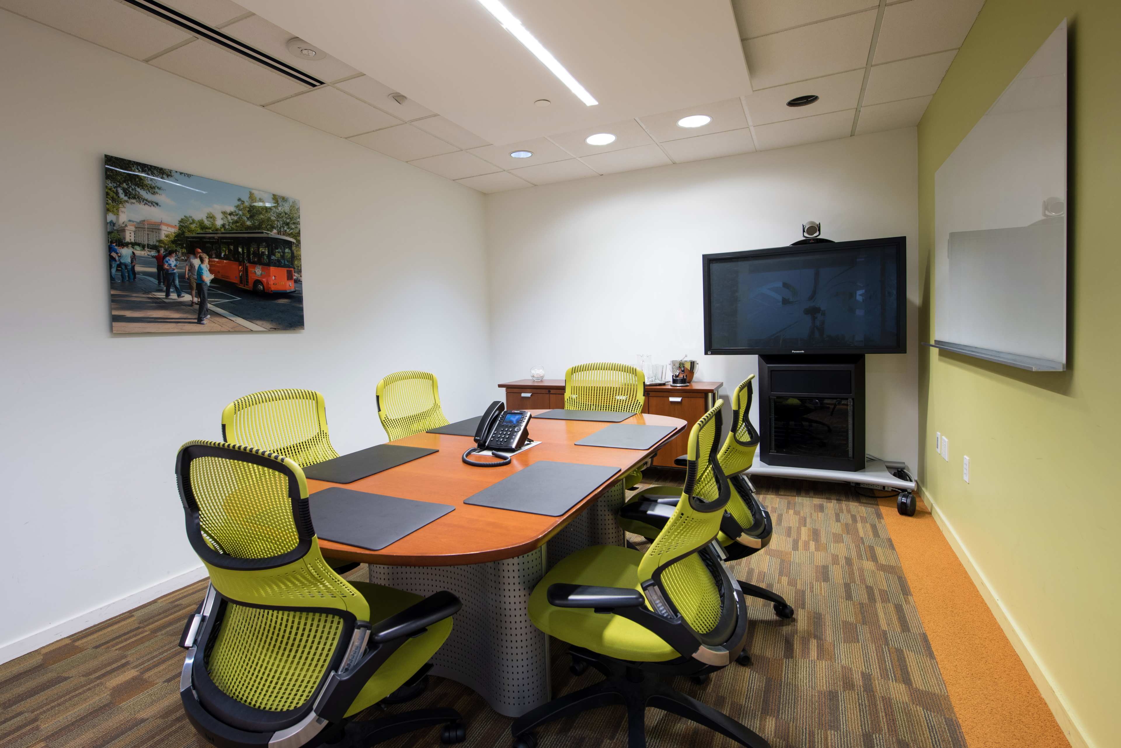 A modern conference room features a rectangular table surrounded by green office chairs, a television mounted on the wall, and a large whiteboard.