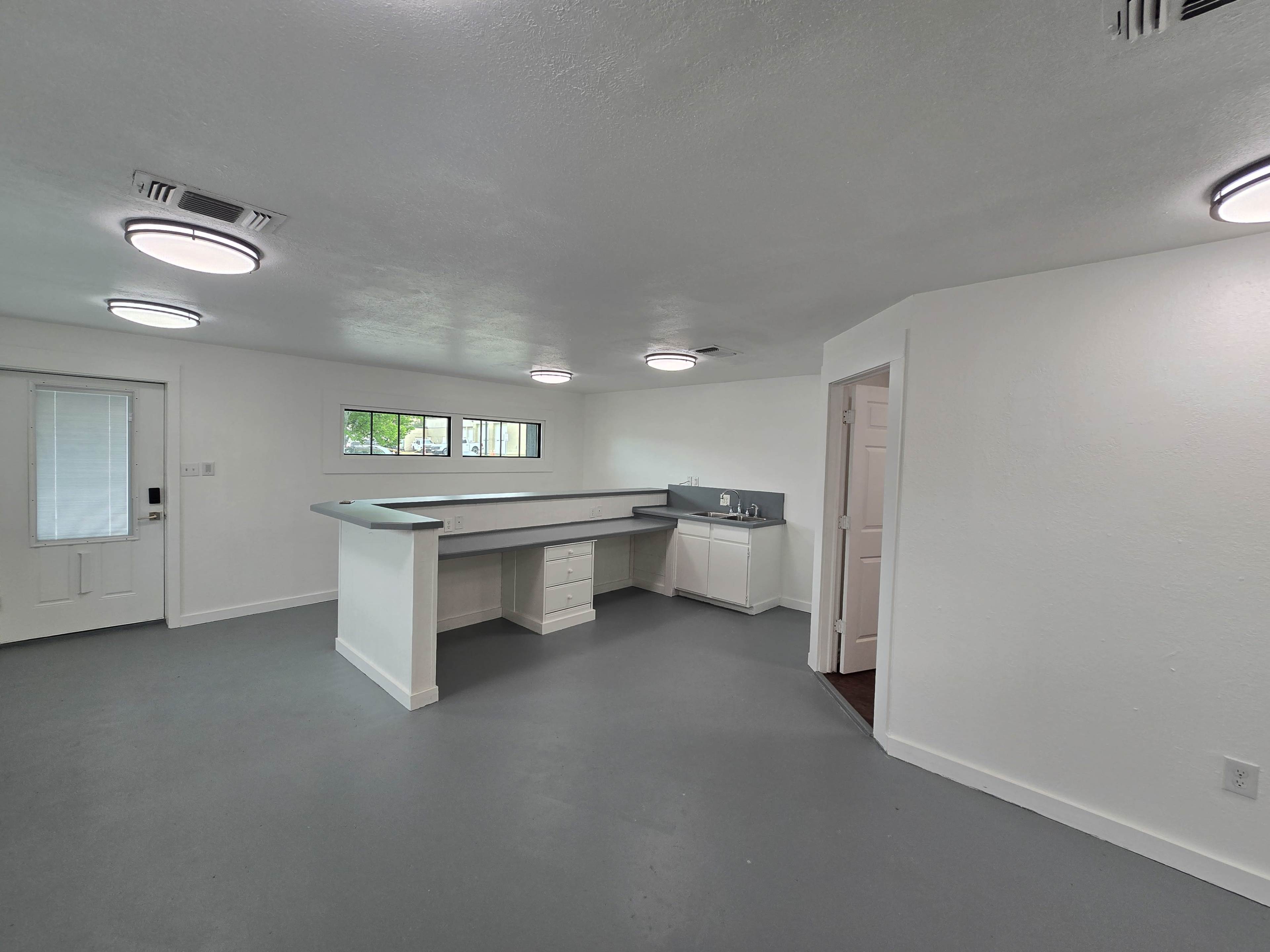 The image shows a spacious, bright room with a gray floor, a kitchen bar area with countertops, and a door leading outside.