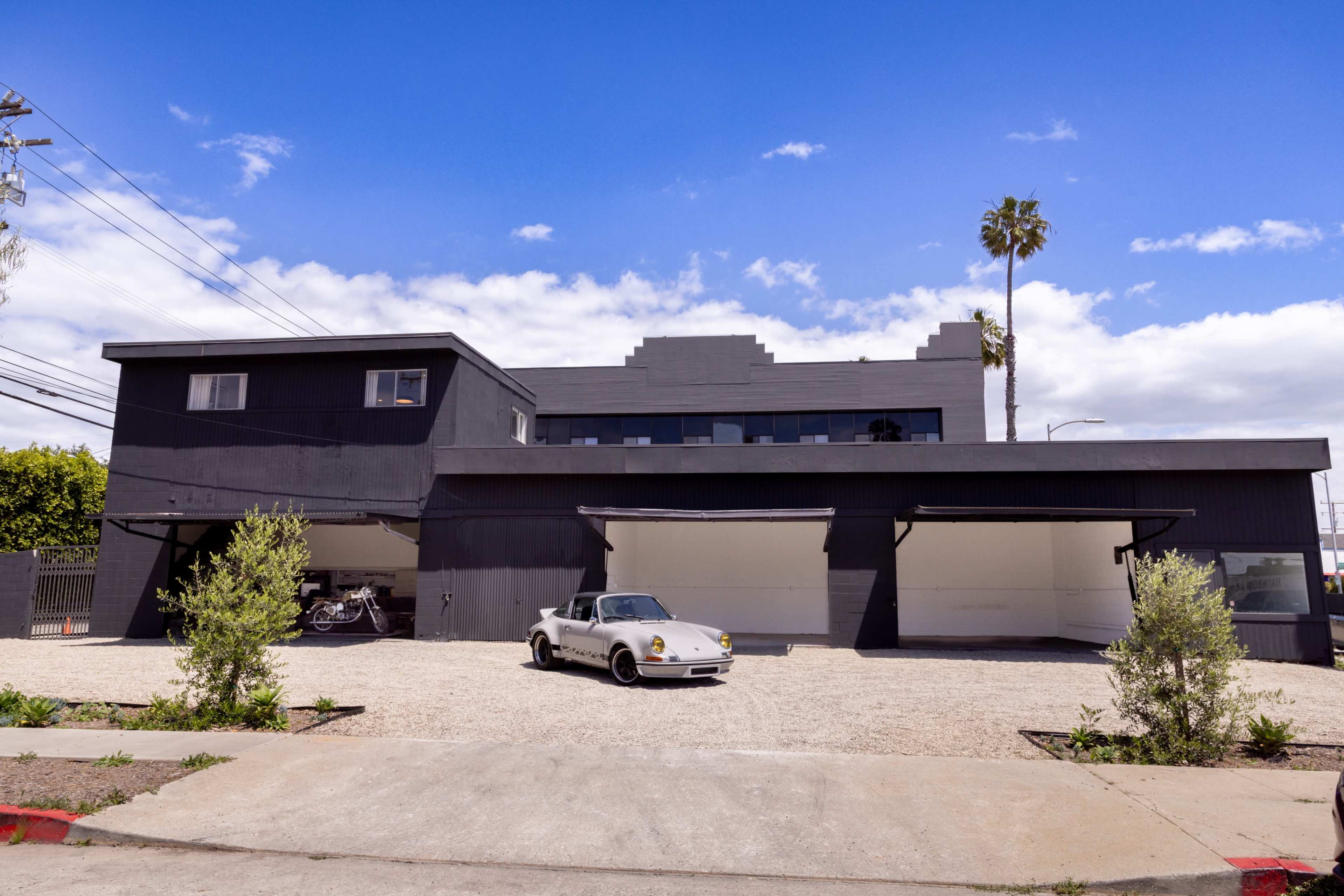 A modern black building with a textured façade features a white sports car parked in front on a gravel area, flanked by small plants and palm trees under a blue sky.