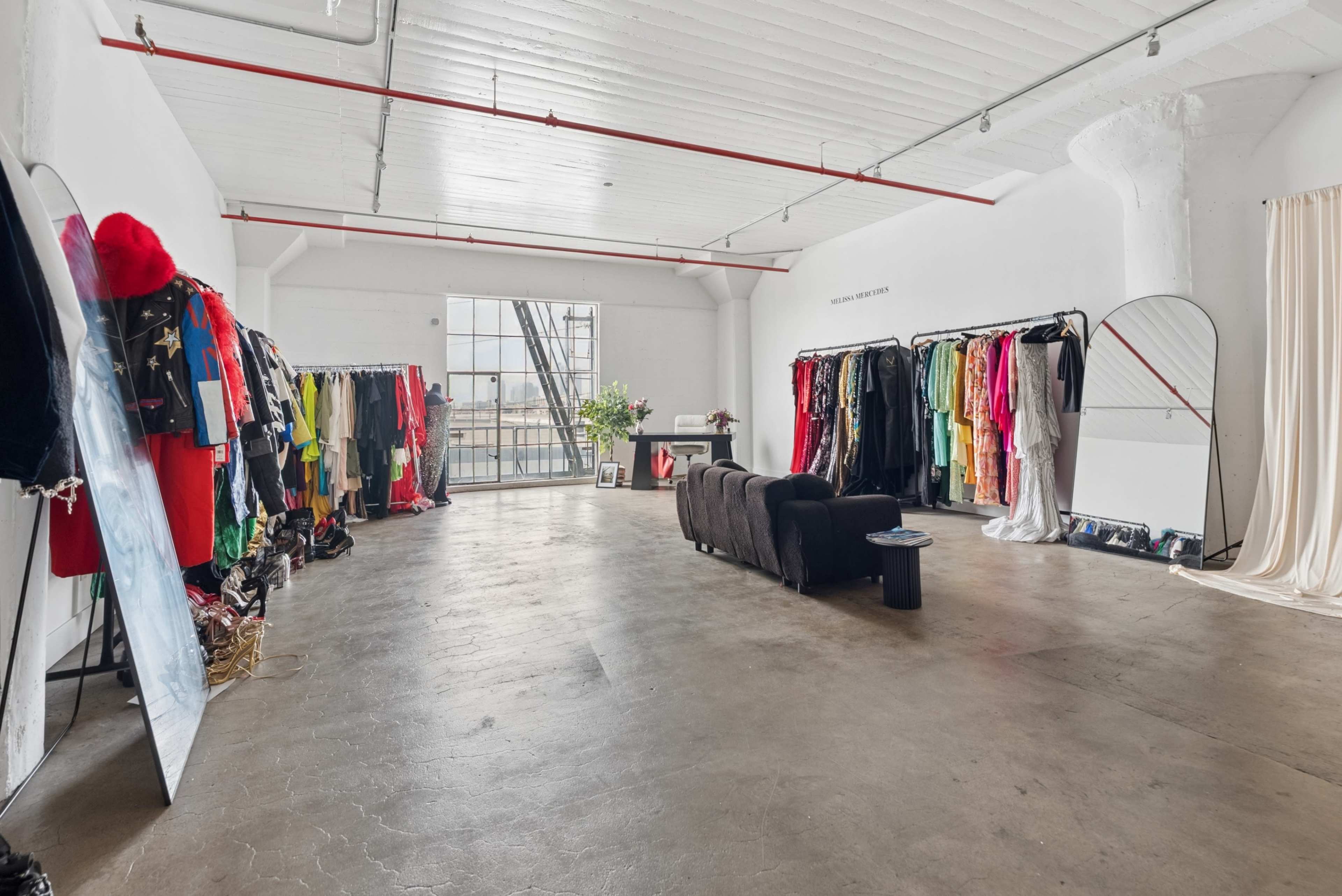 The image shows a spacious, well-lit room with racks of colorful clothing on one side and a black couch and mirror on the opposite side.