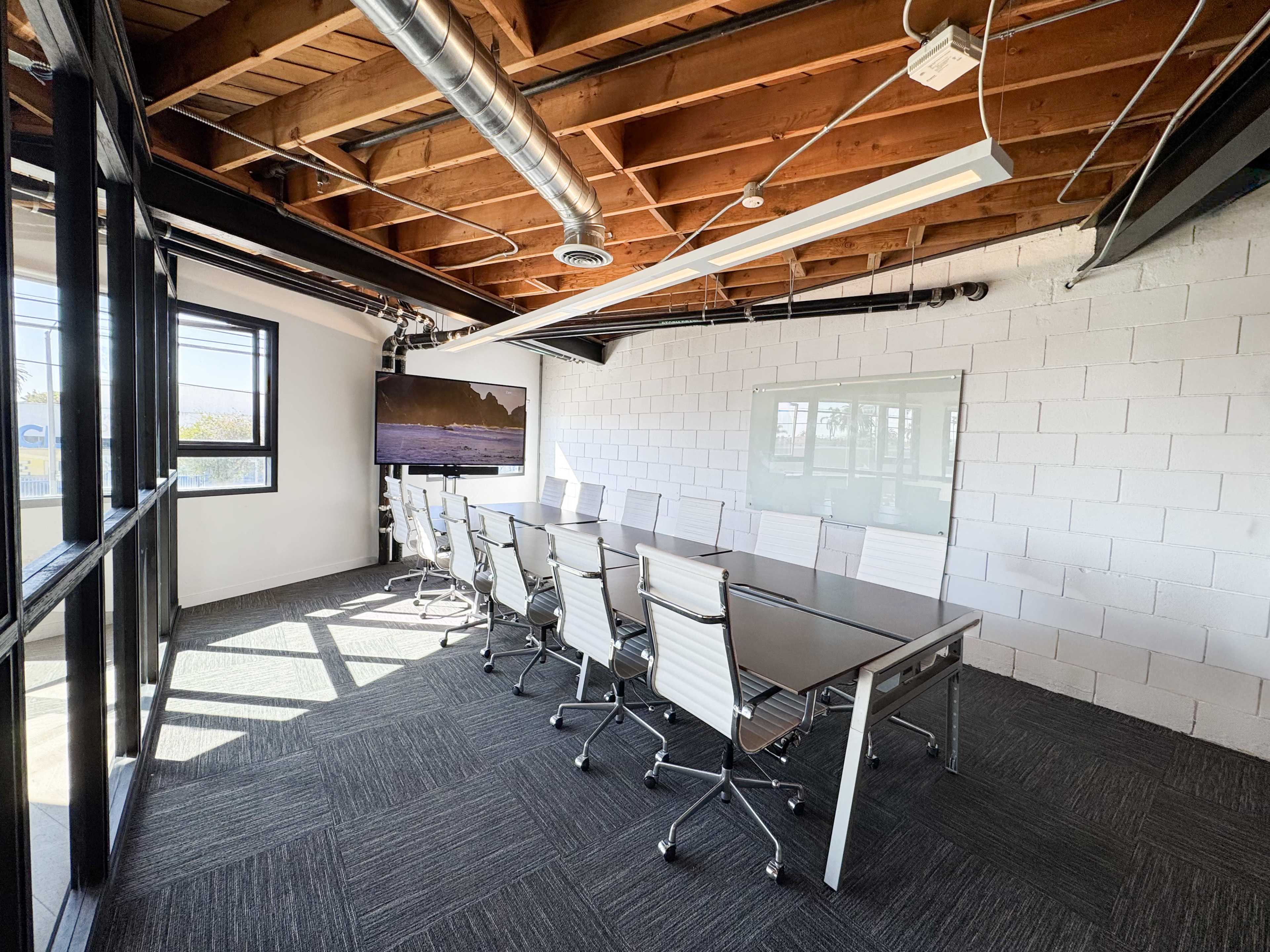The image shows a modern conference room featuring a long table, ergonomic chairs, and large windows, with a presentation screen on one wall.