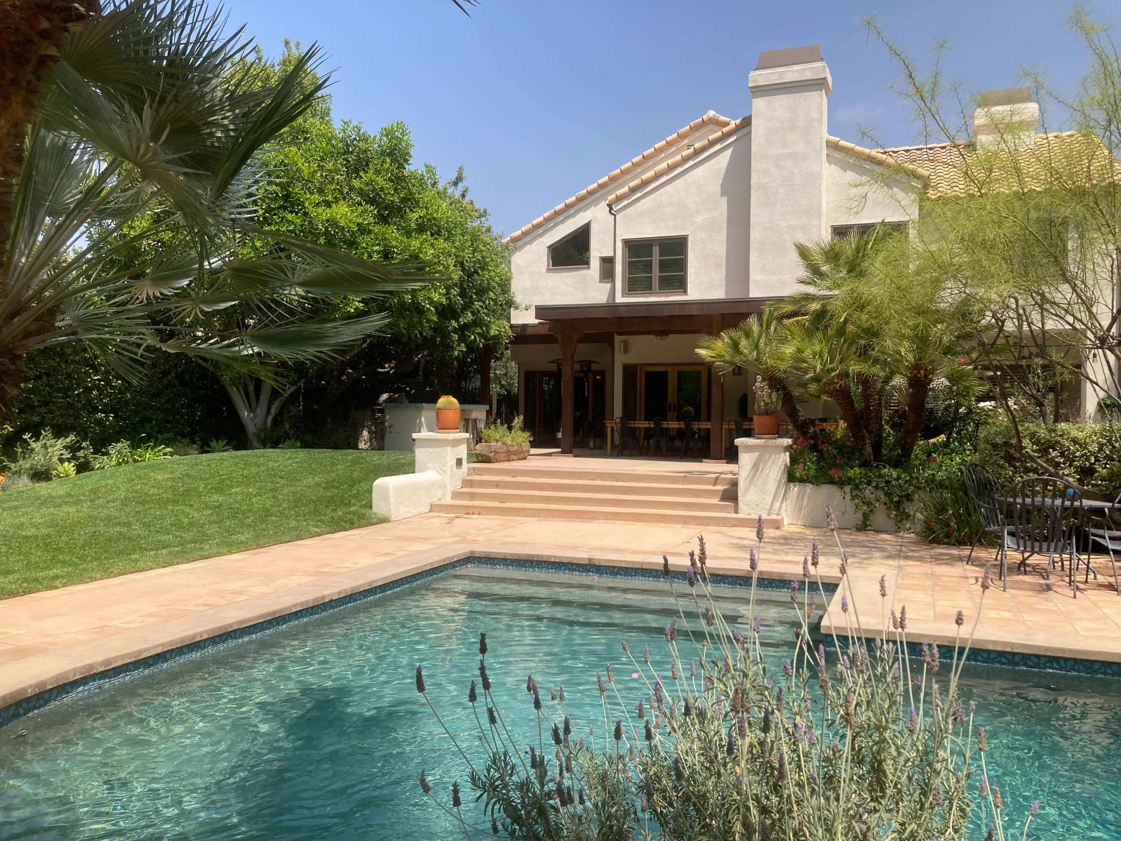A two-story house with a tiled roof and a patio overlooks a swimming pool surrounded by greenery.