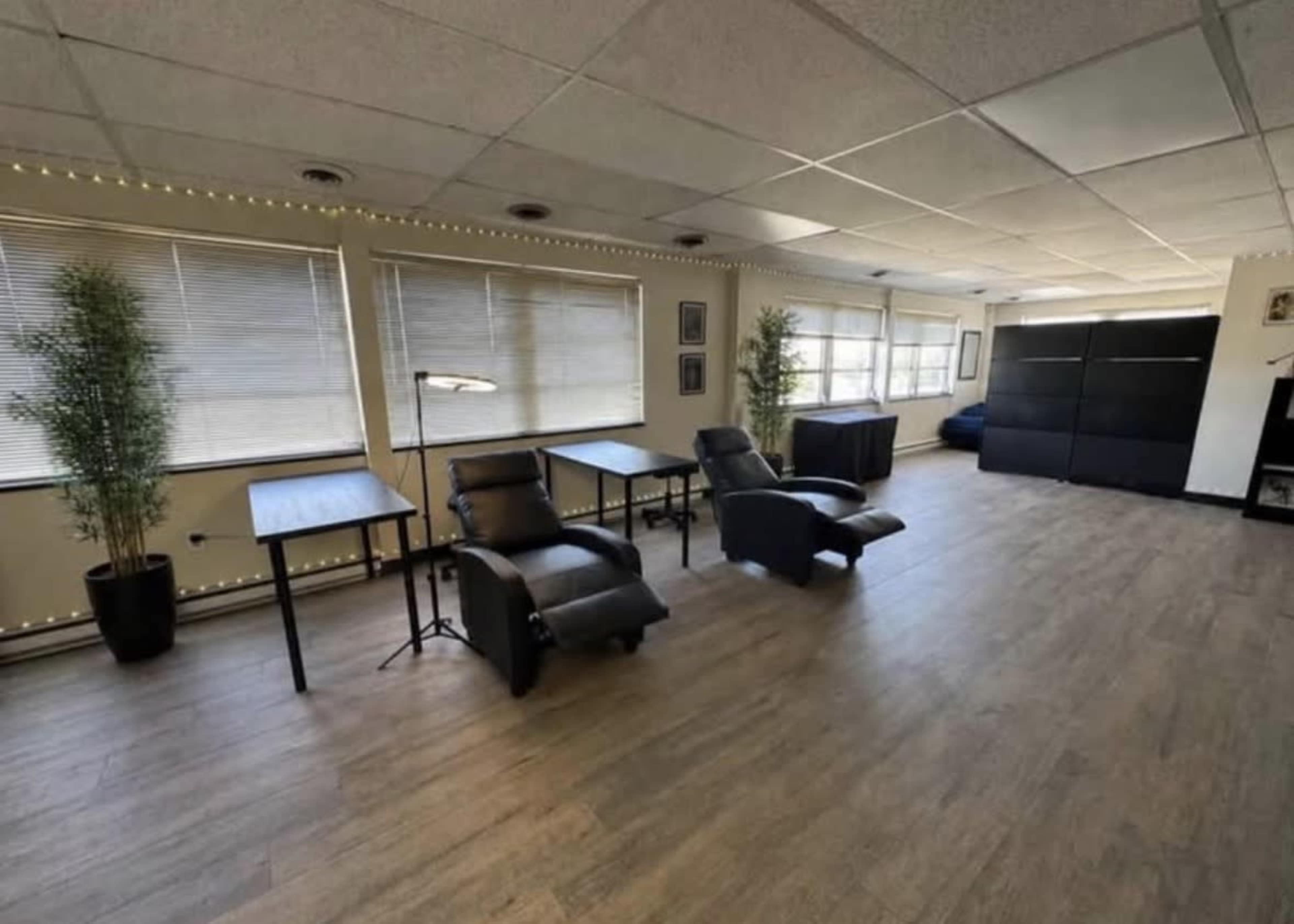 The image shows a spacious room with two reclining chairs, two tables, and large windows lined with plants, accompanied by a dark couch and shelving unit against the wall.