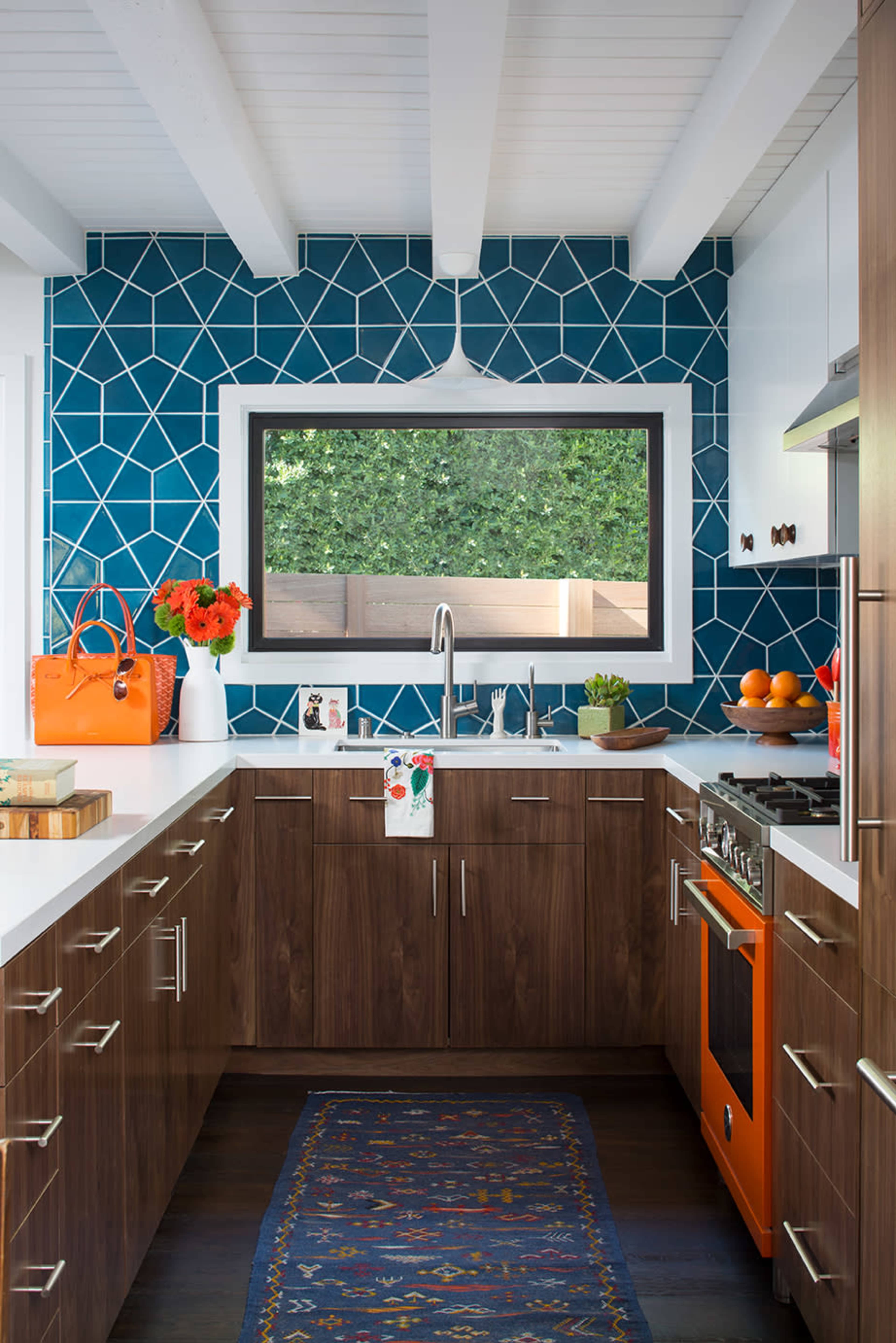 A modern kitchen features a bold geometric blue tile backsplash, wooden cabinetry, and orange accents, including an oven and a handbag.