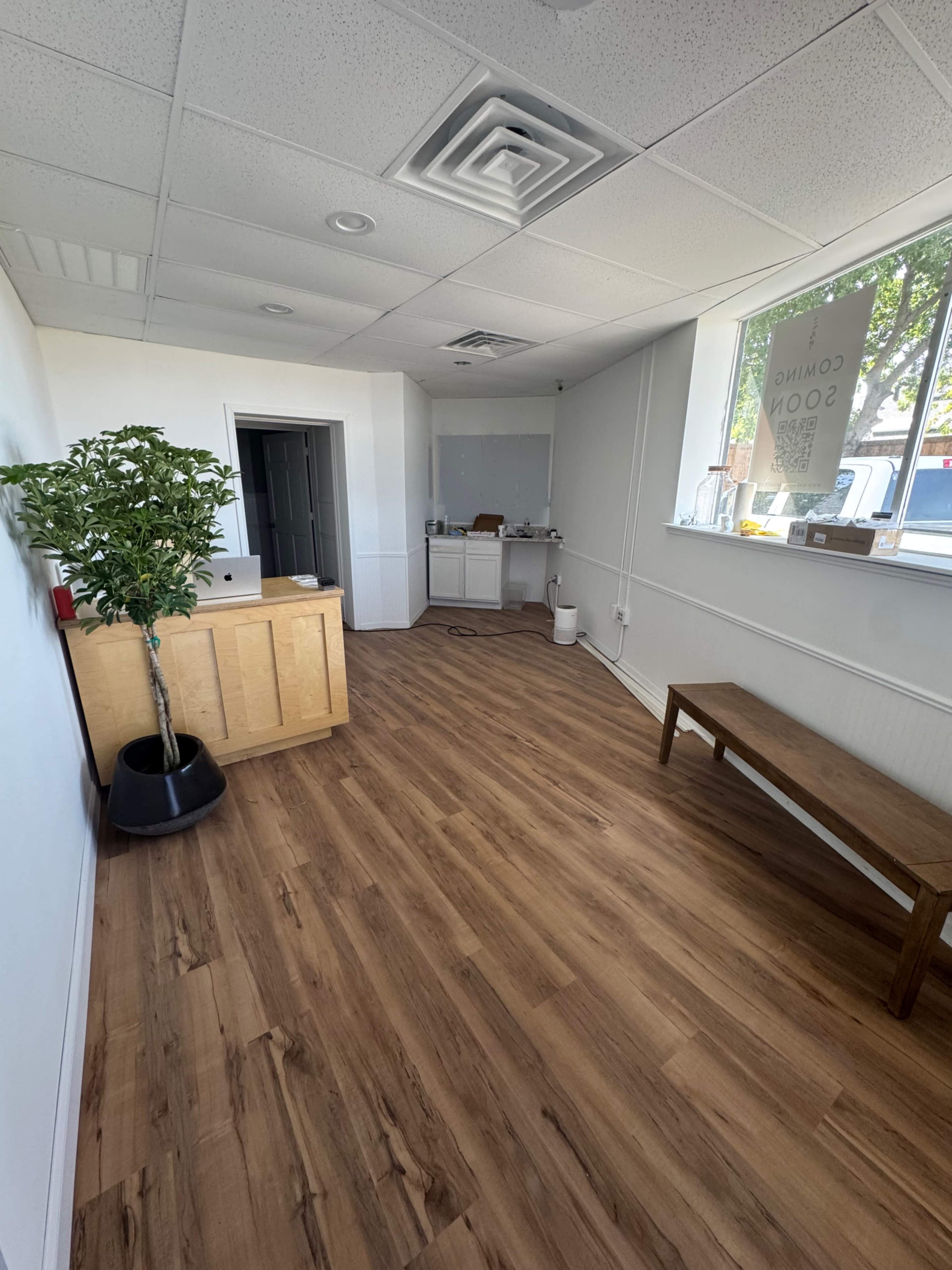 The image shows an empty office space with wooden flooring, a reception desk, a bench, and a large window.