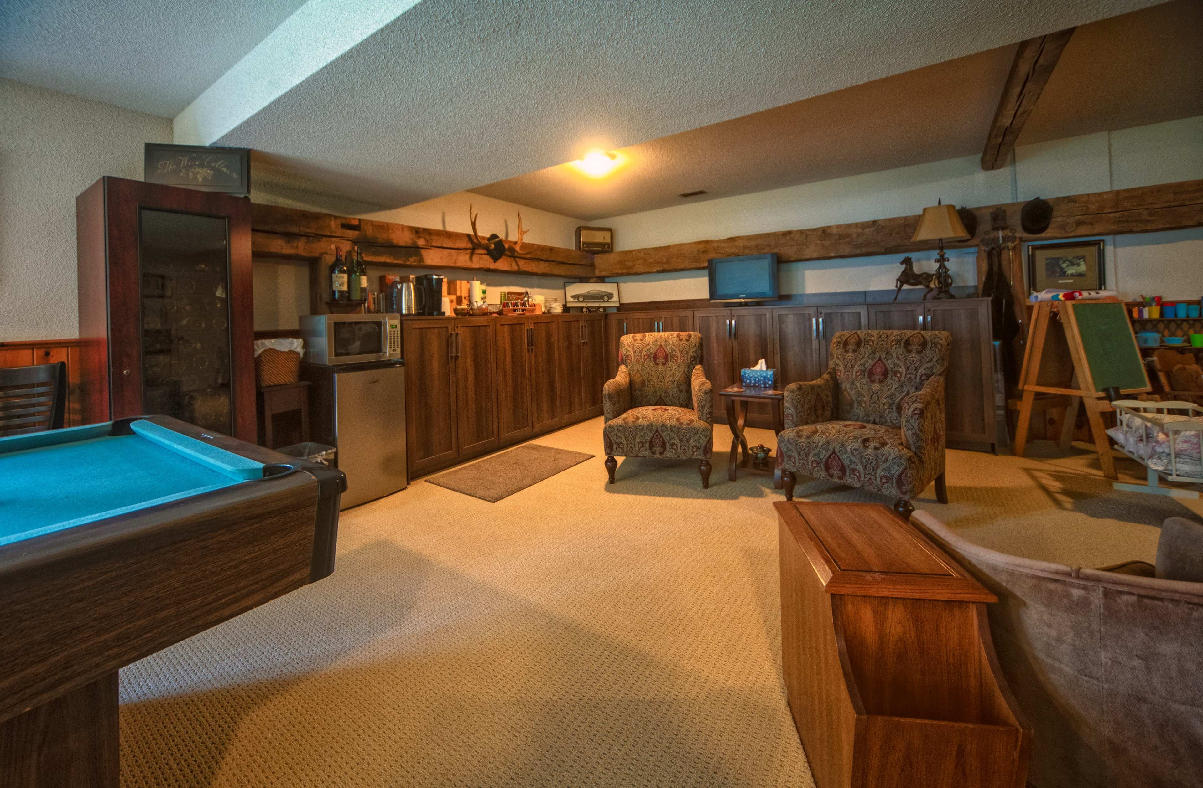 The image shows a finished basement featuring a pool table, two patterned armchairs, a small kitchen area with appliances, and various pieces of furniture arranged in a cozy living space.