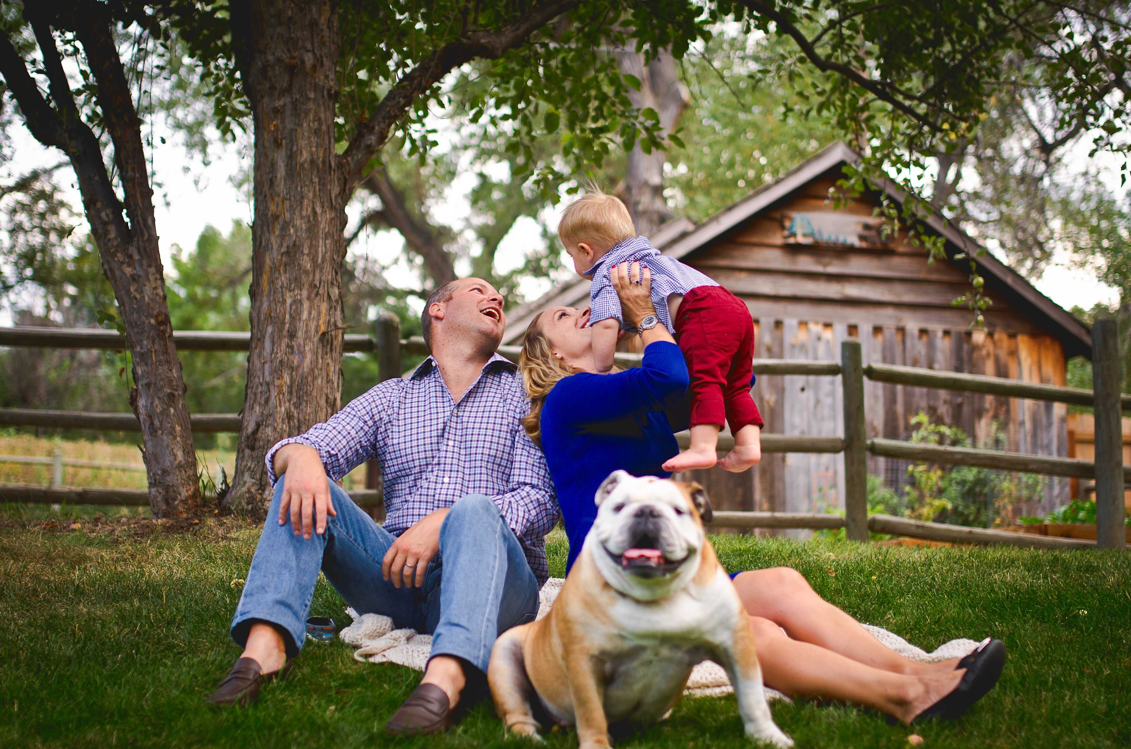 A family sits on the grass in a yard, with a baby being lifted by a woman, while a dog rests nearby and a wooden shed is in the background.