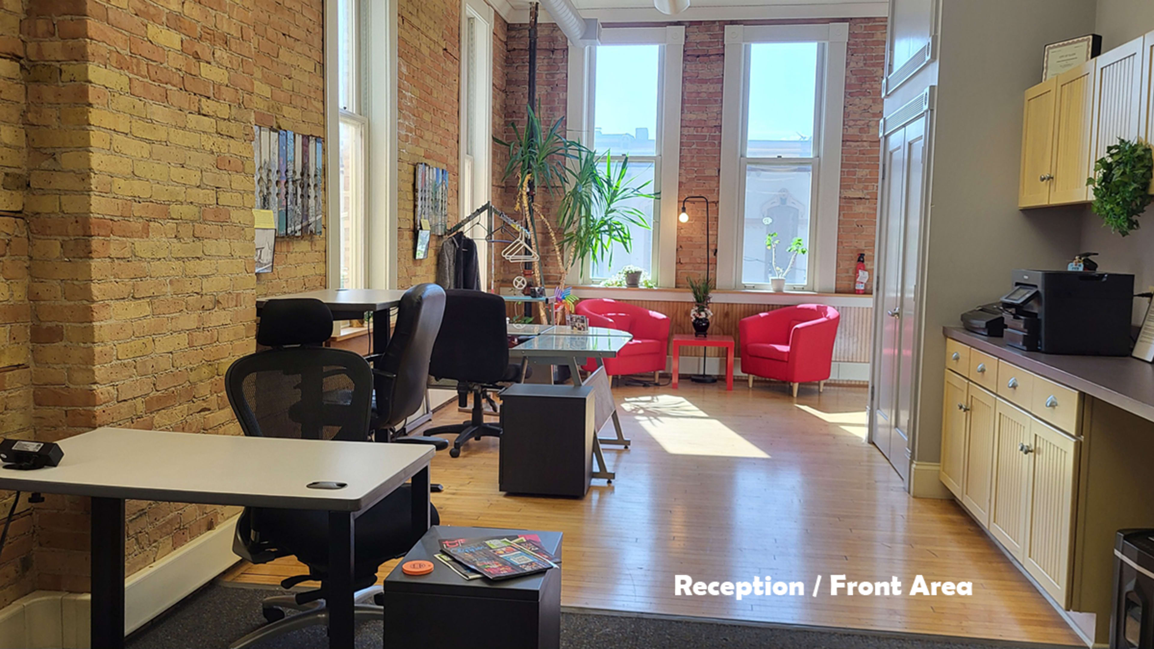 The image shows a reception area with exposed brick walls, two red chairs, a desk with a chair, and a window letting in natural light.