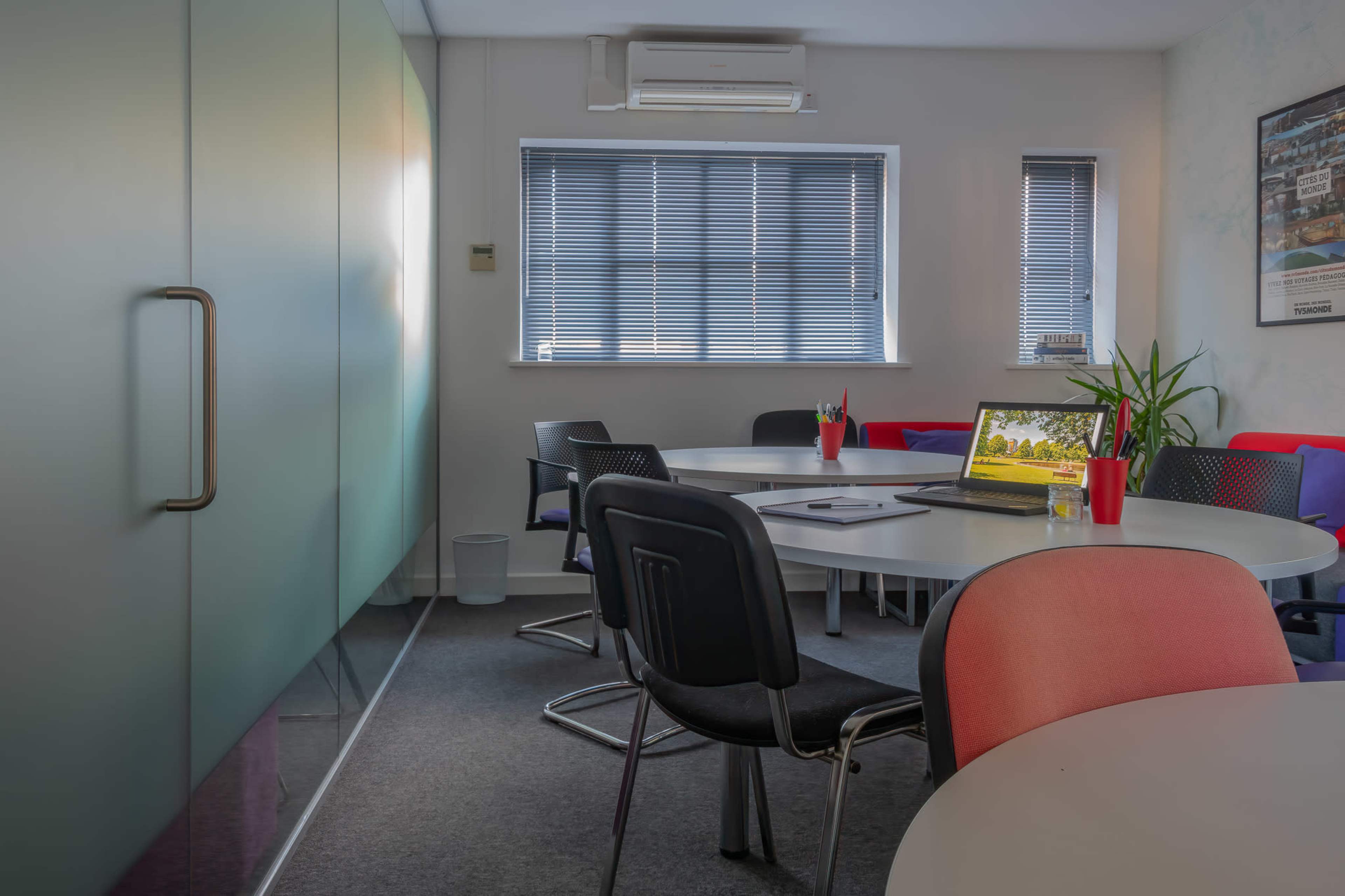 A modern conference room features a round table surrounded by black and red chairs, with a laptop open on the table and large windows partially covered by blinds.