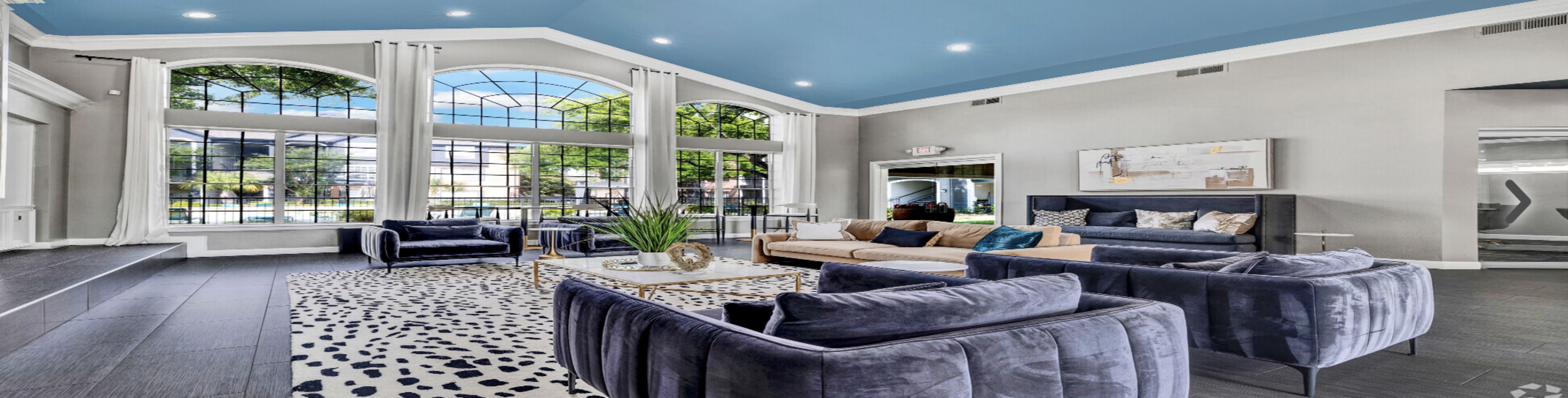 The image shows a spacious living area with large windows, modern furniture, and a decorative rug.