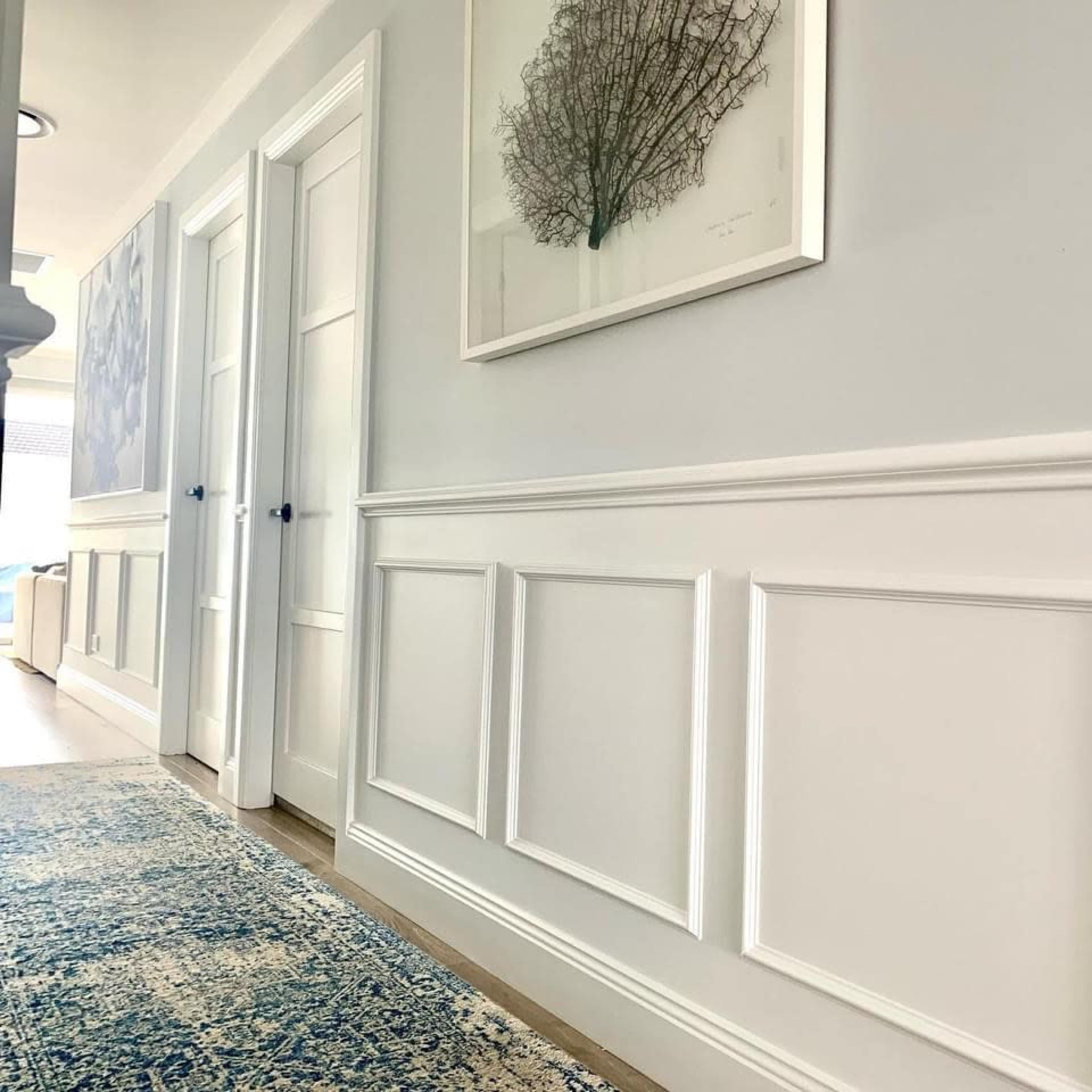 A hallway with white-paneled walls, two closed doors, and a framed artwork of a tree on the wall, accompanied by a blue area rug.