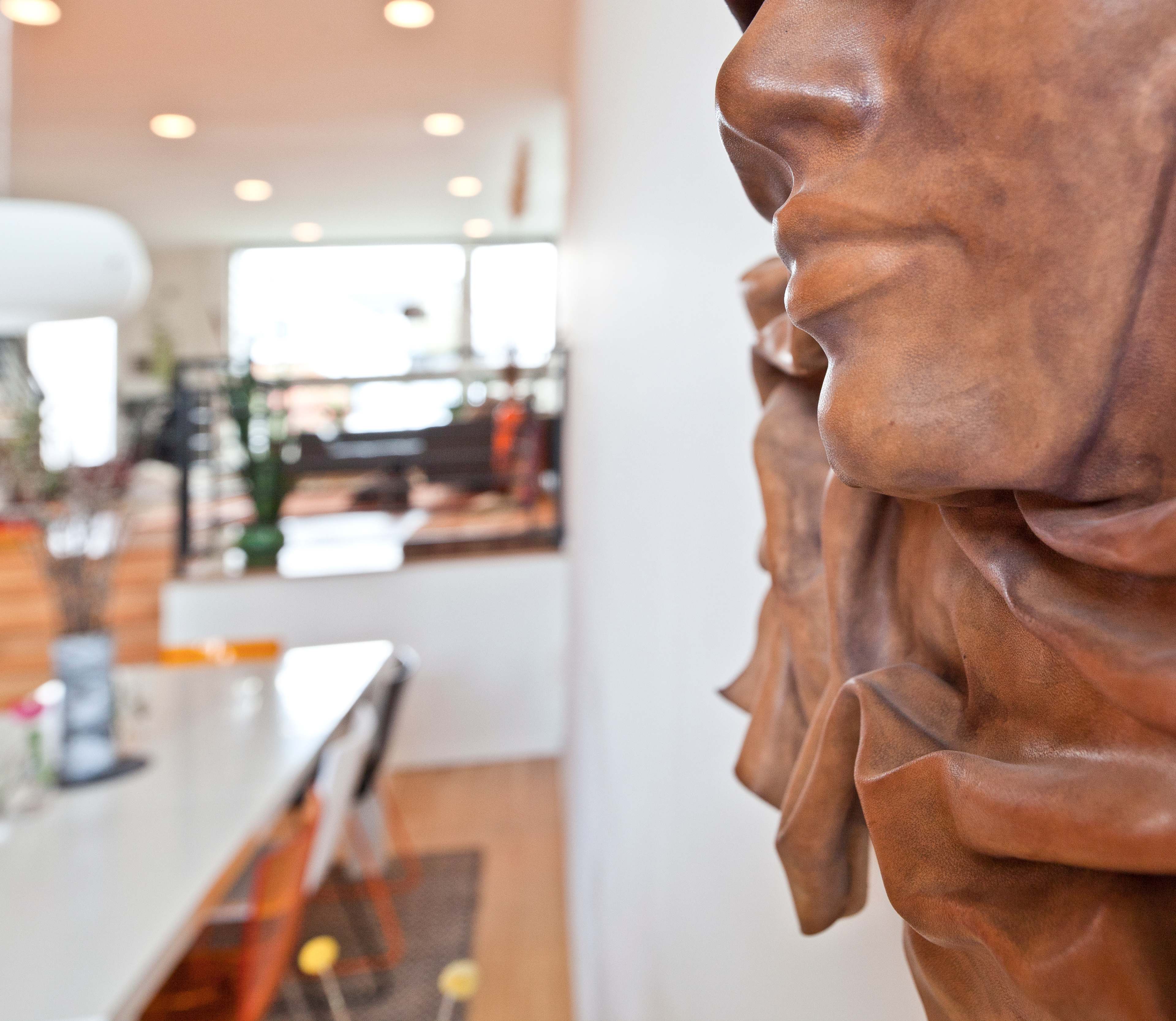 A textured sculpture of faces is mounted on a wall, with a dining area and large windows visible in the background.
