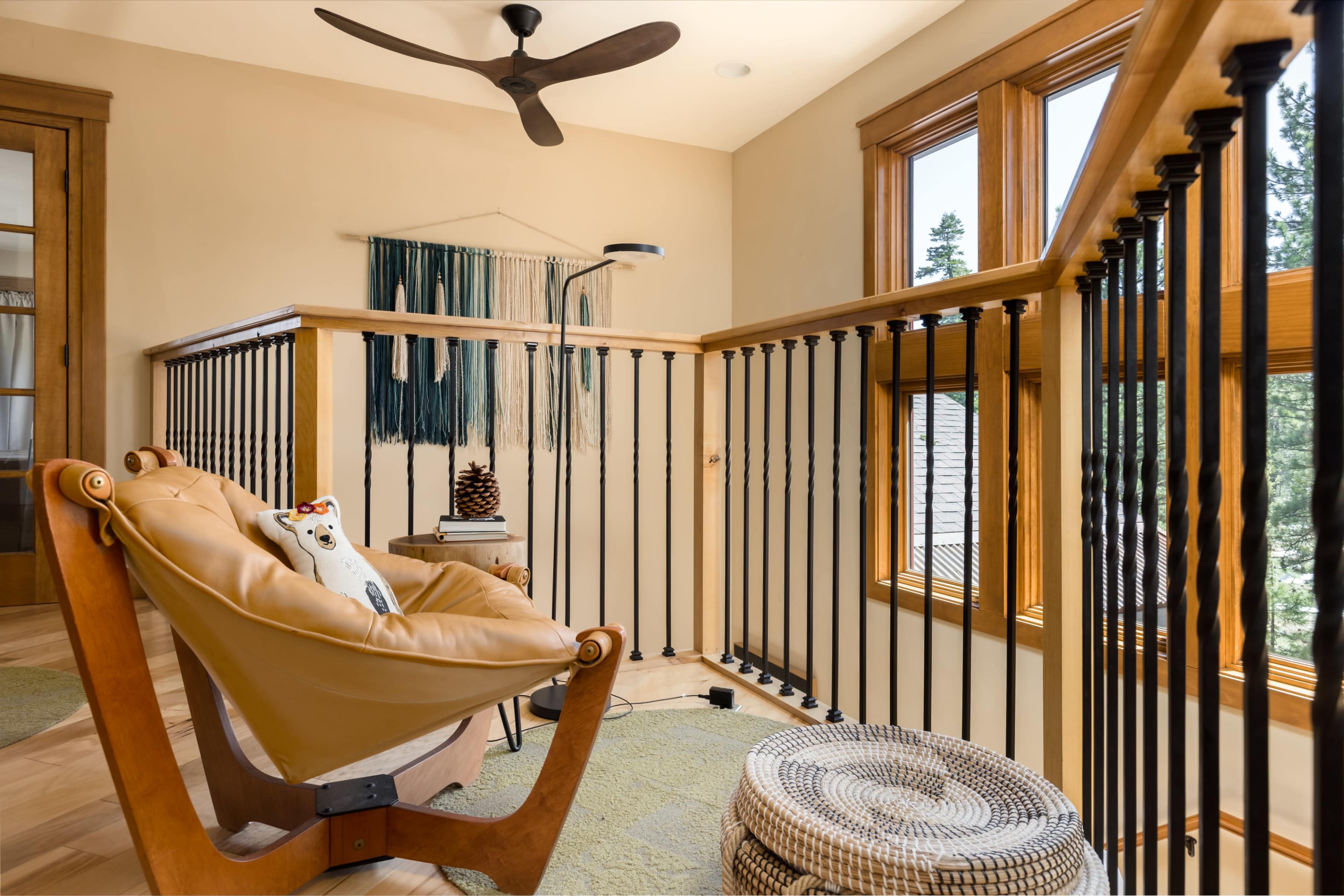 The image shows a sitting area on a second floor, featuring a modern chair, a woven wall hanging, and large windows providing natural light.