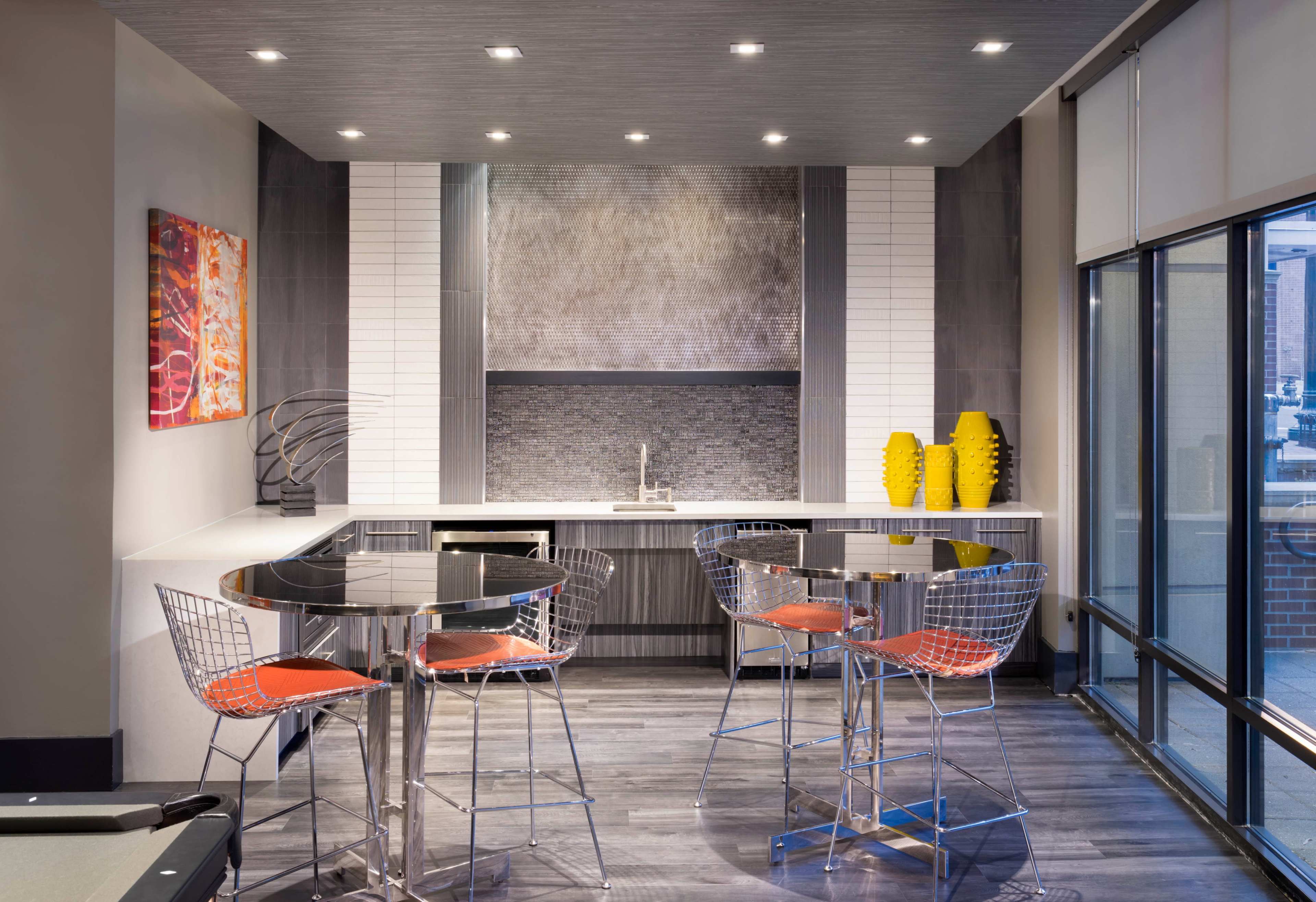 A modern kitchen area with a bar-style dining set, featuring chrome stools and a sleek countertop, complemented by decorative yellow vases and wall art.