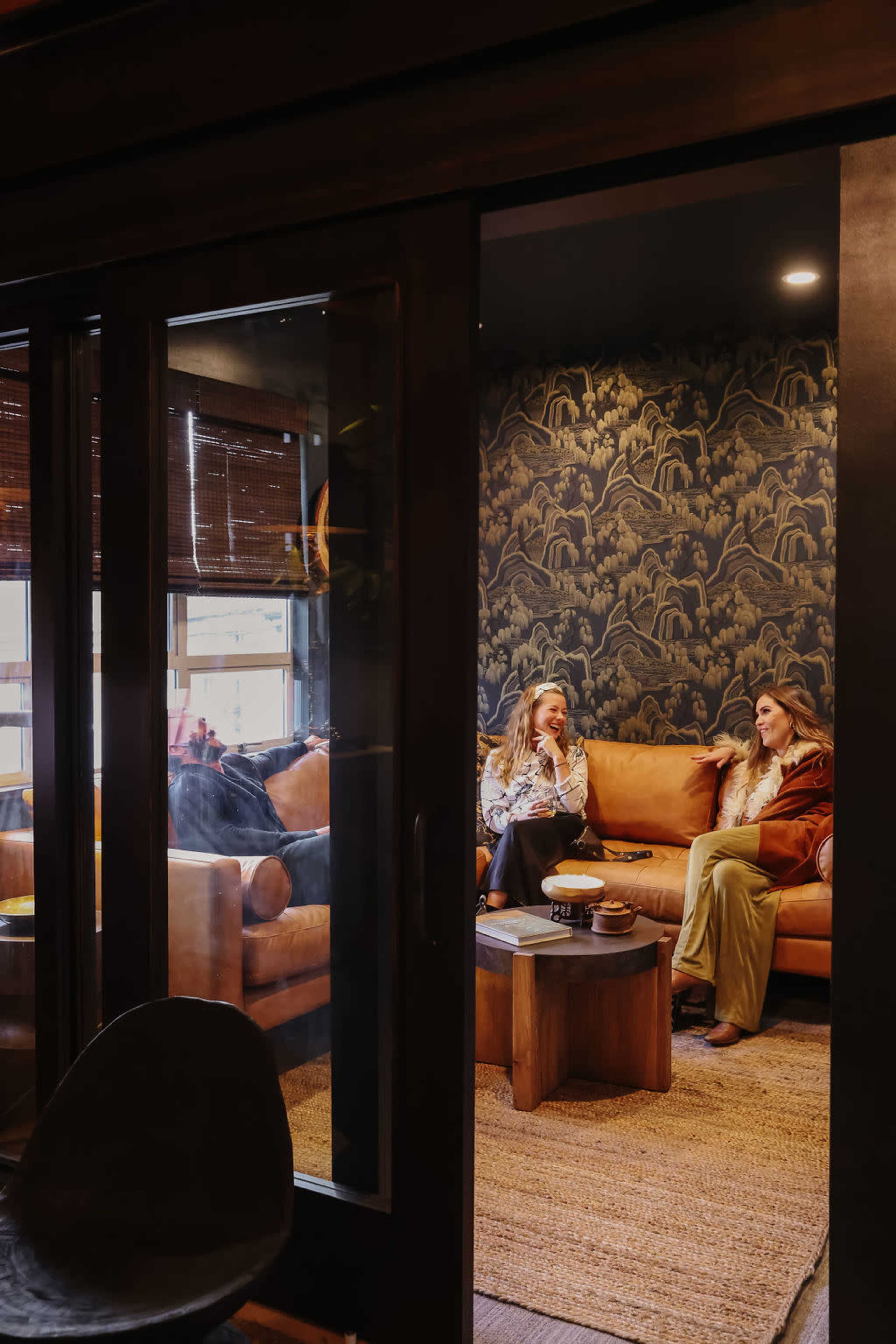 Two women are seated on a sofa in a cozy room with a dark, patterned wallpaper, engaged in conversation.