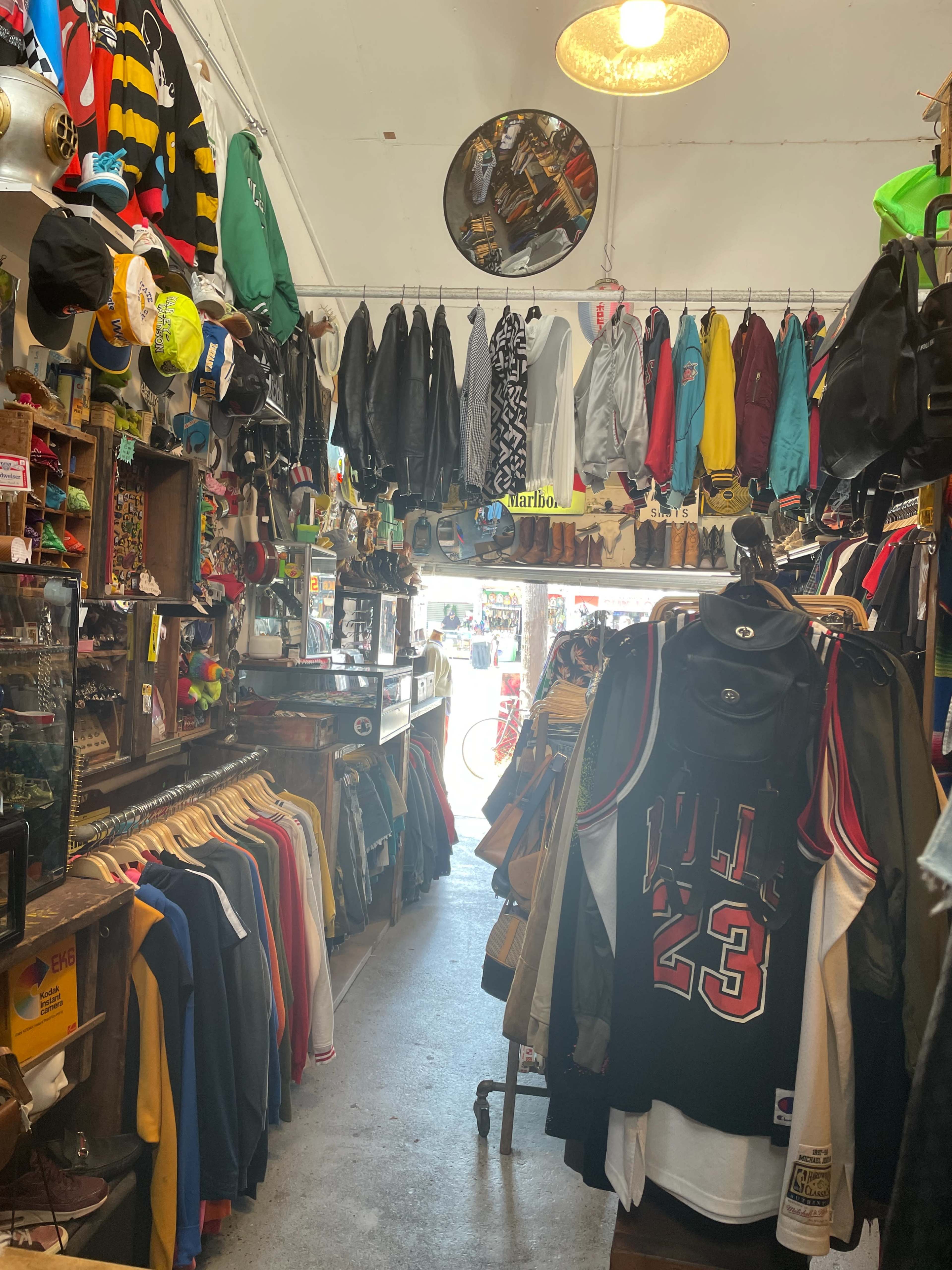 The image shows a narrow vintage clothing store aisle lined with various jackets and shirts hung from wooden racks and walls, while colorful apparel is draped from the ceiling.