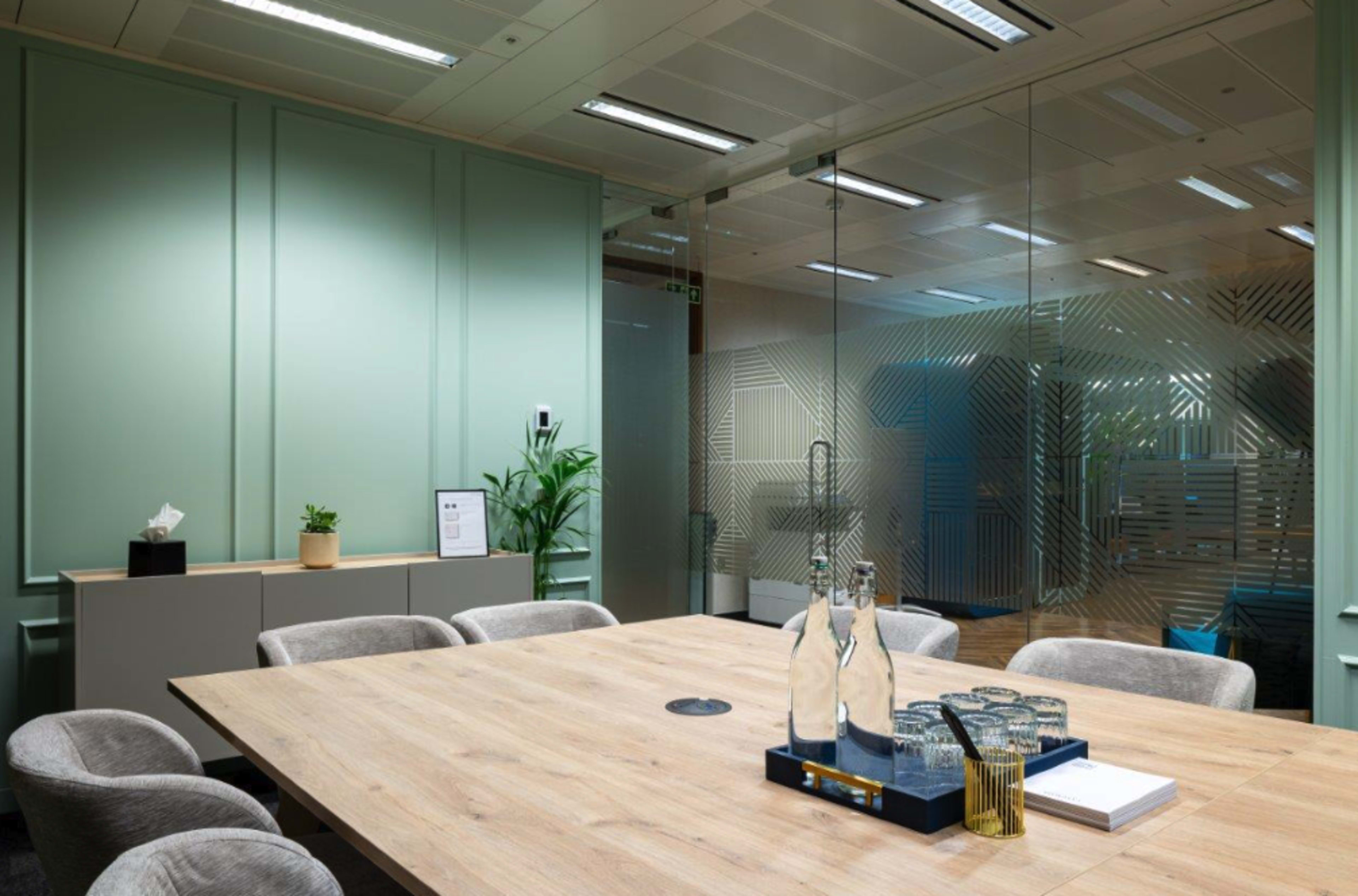 A modern conference room features a wooden table surrounded by upholstered chairs, with glass walls and a small plant in the corner.