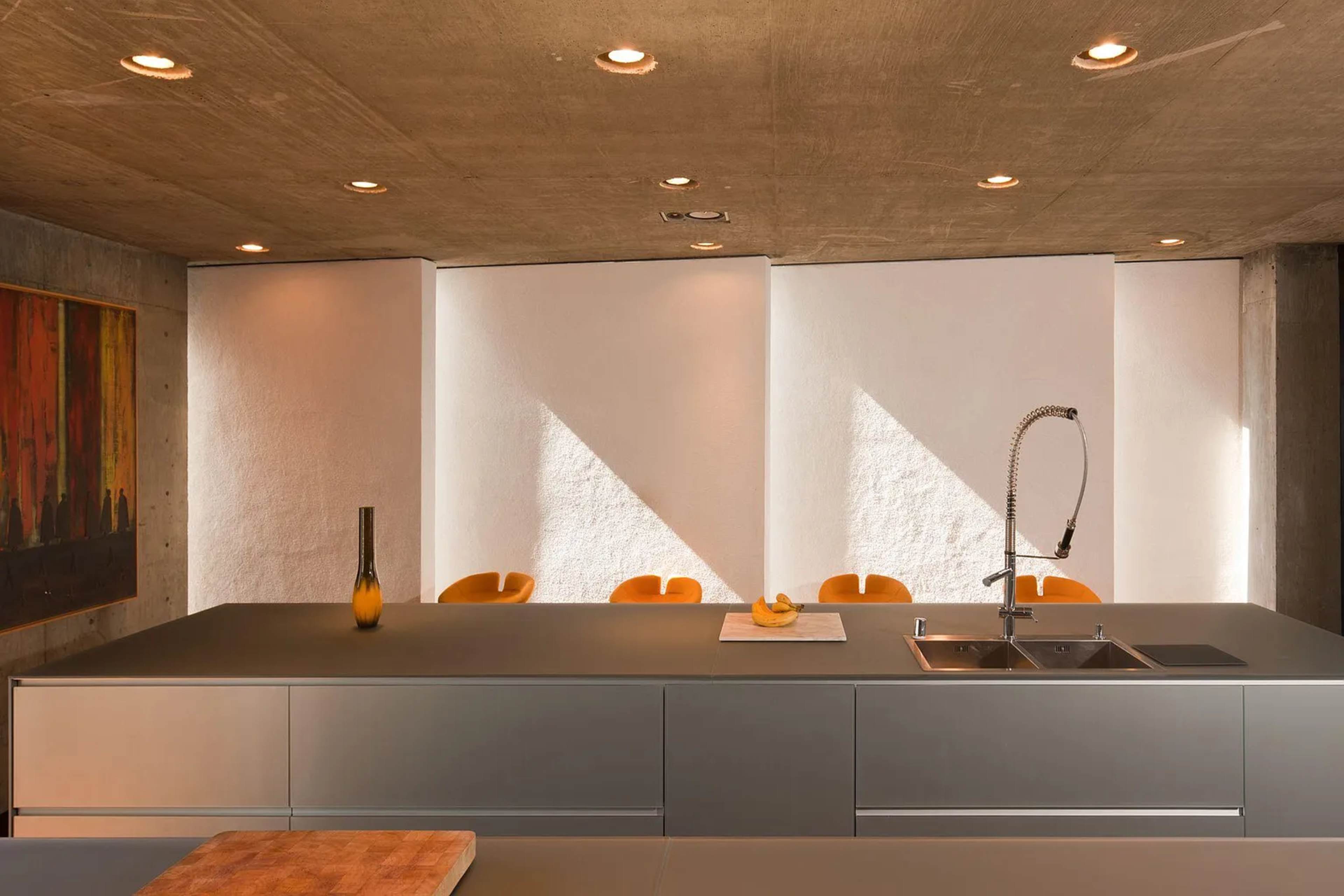 A modern kitchen features a concrete ceiling, minimalist cabinetry, and bright yellow chairs around a table, with light casting geometric shadows across the space.