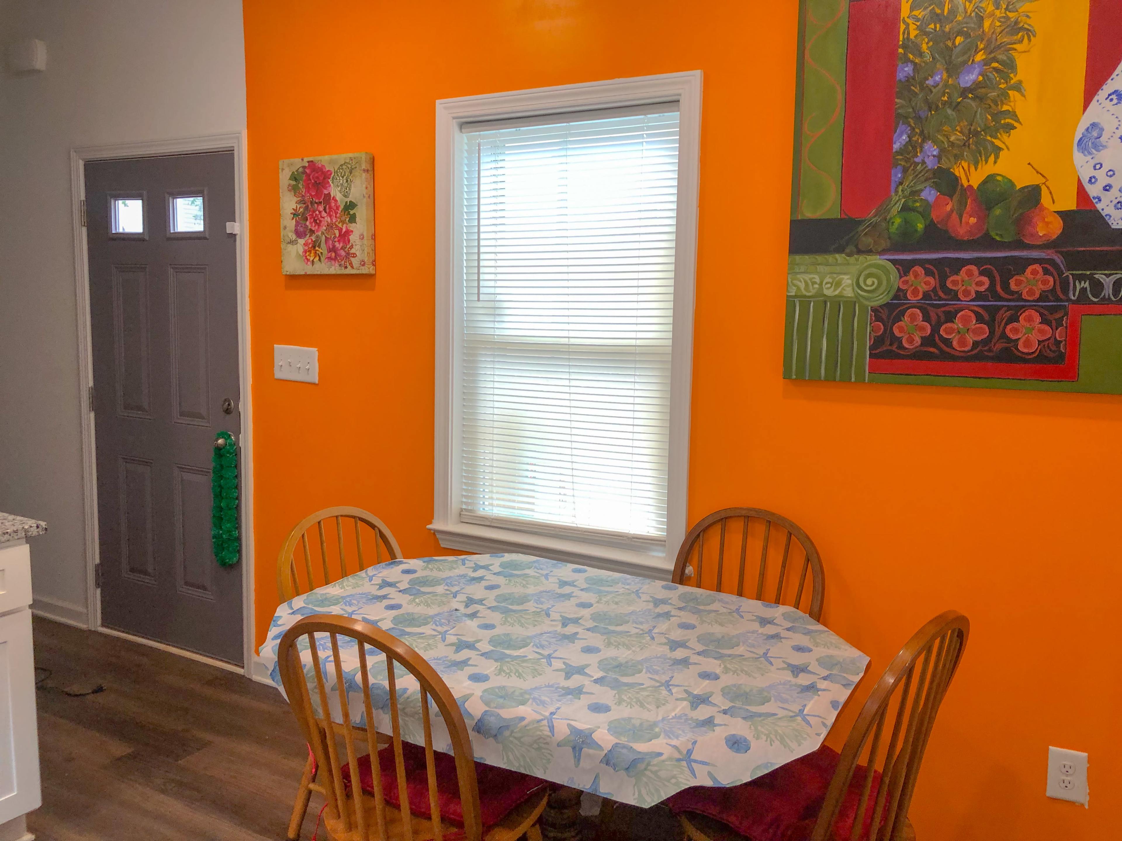 A wooden table surrounded by four chairs sits in a brightly colored orange dining area, with a window and a door in the background.