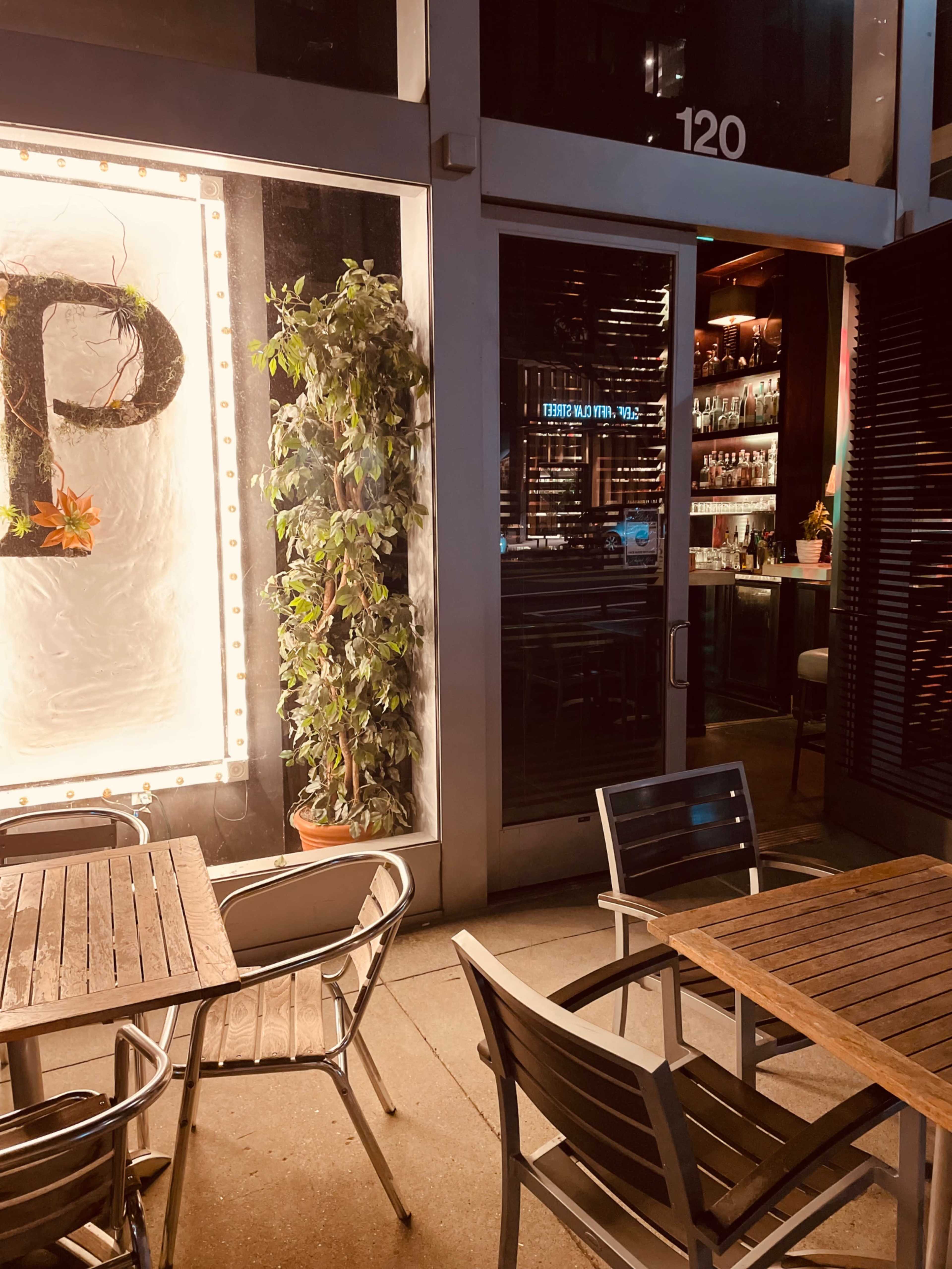 An outdoor dining area features metal chairs and wooden tables beside a brightly lit bar visible through large glass windows.
