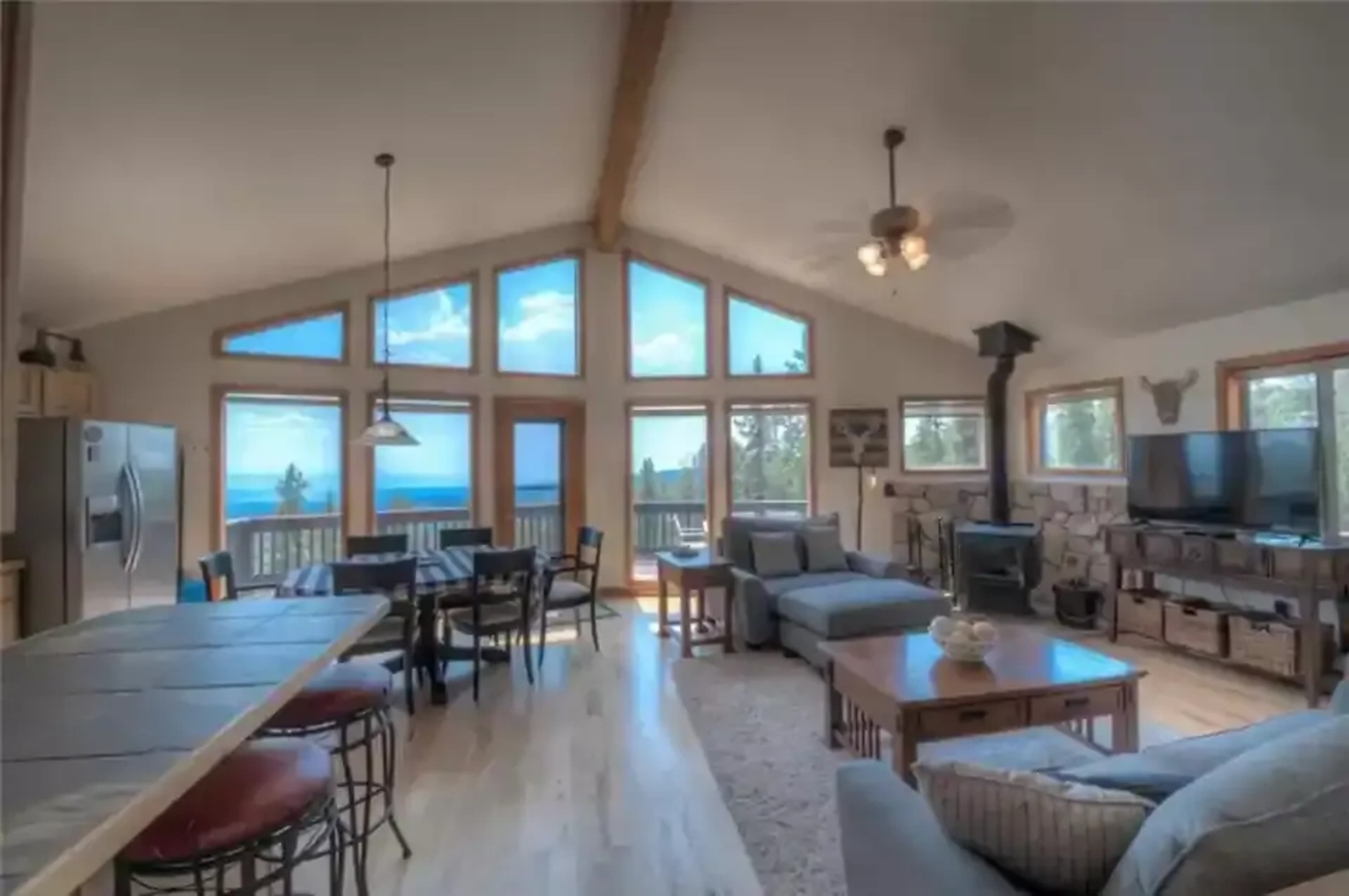 A spacious living area with large windows offering a view of the outdoors, featuring a dining table, kitchen, and comfortable seating.