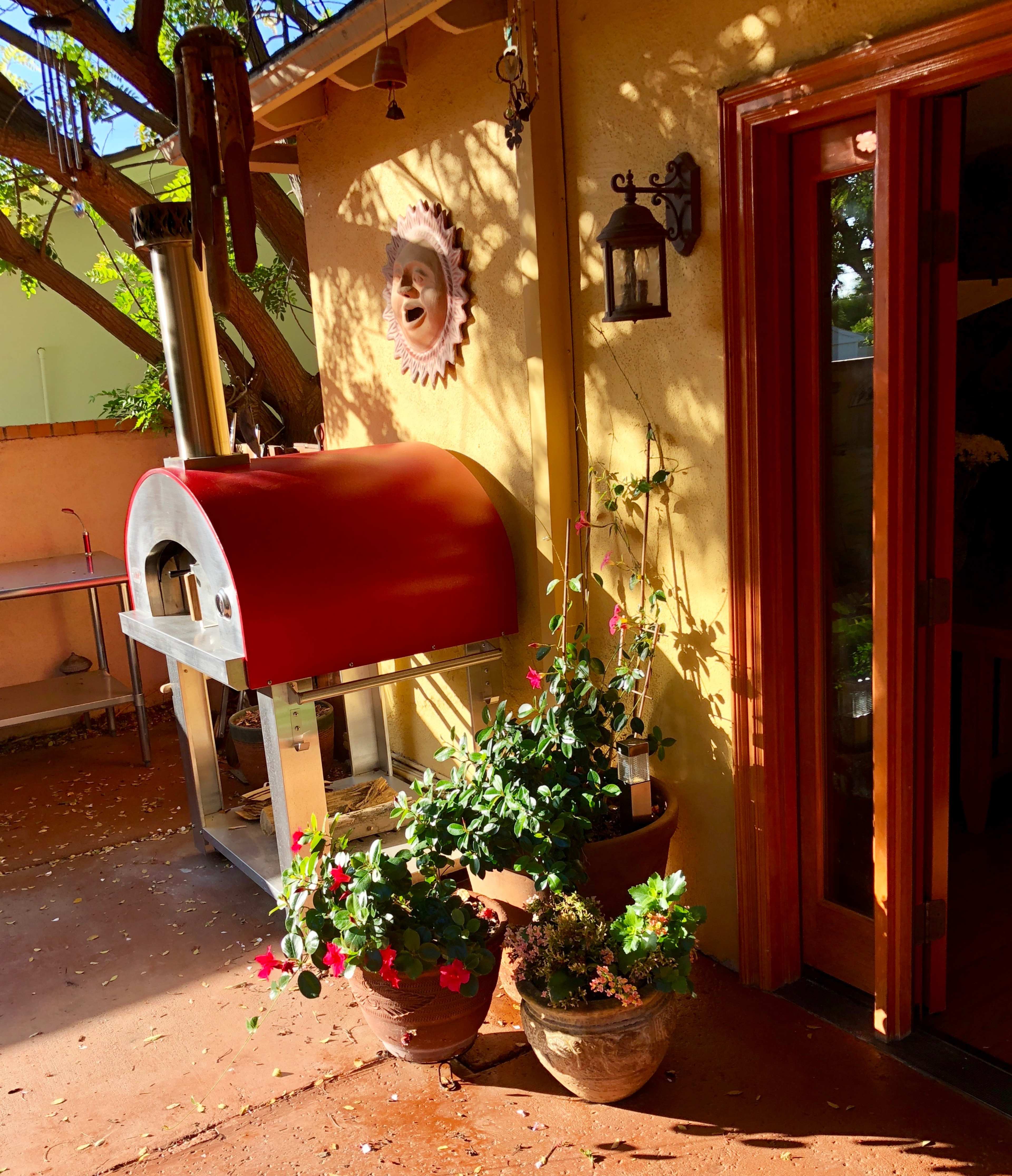 The image shows a red outdoor pizza oven next to potted plants against a sunlit wall, with a wooden door partially open to an interior space.