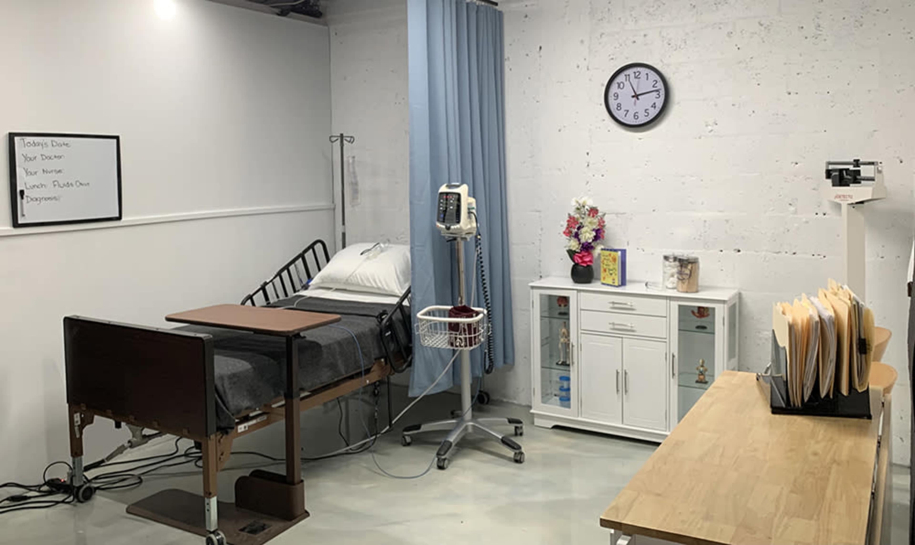 The image shows a medical examination room with a hospital bed, a vital signs monitor, and a clock on the wall, along with a small cabinet and a table covered with file folders.