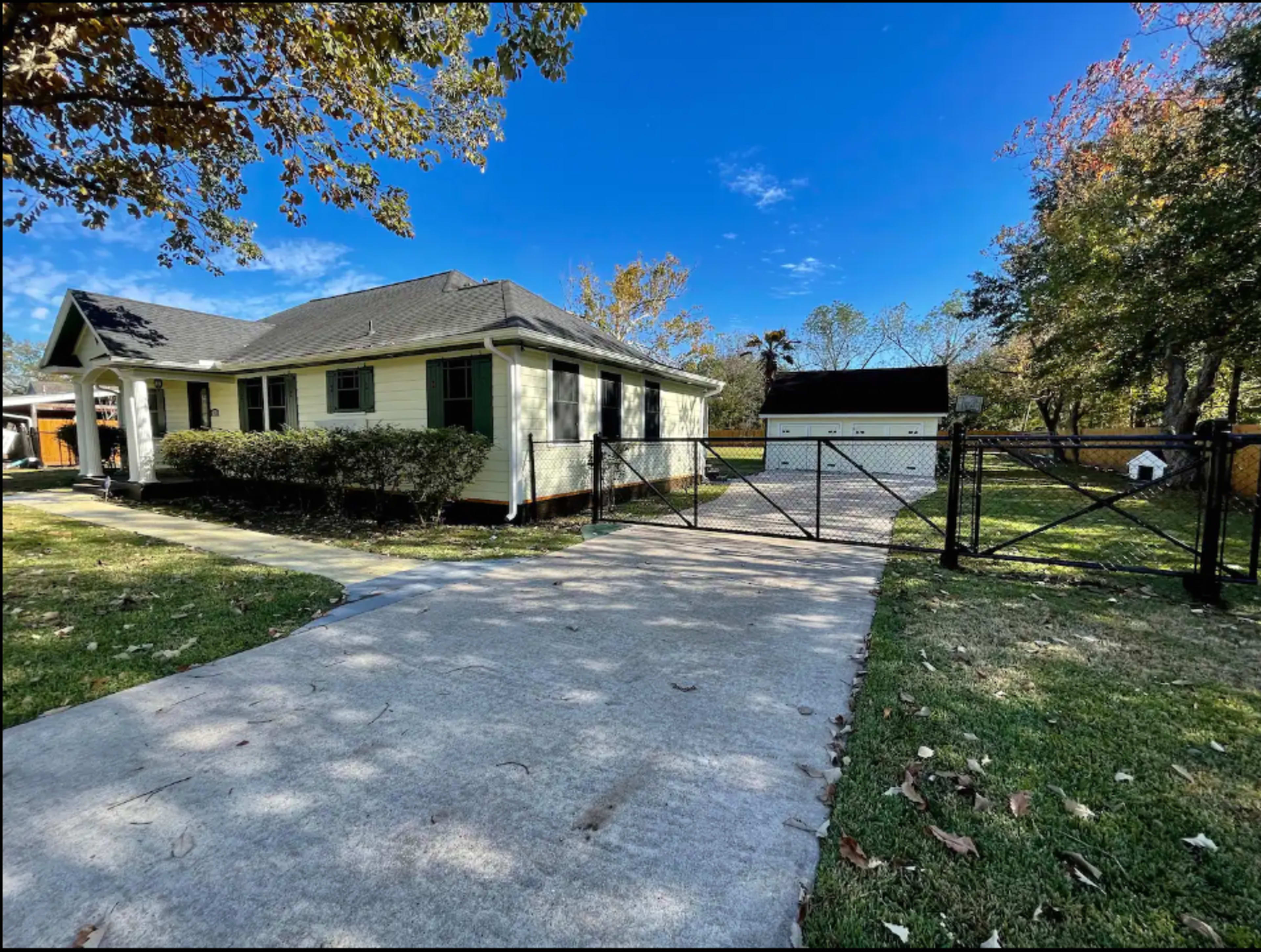 A single-story yellow house with green shutters and a paved driveway leads to a fenced area with a detached garage.
