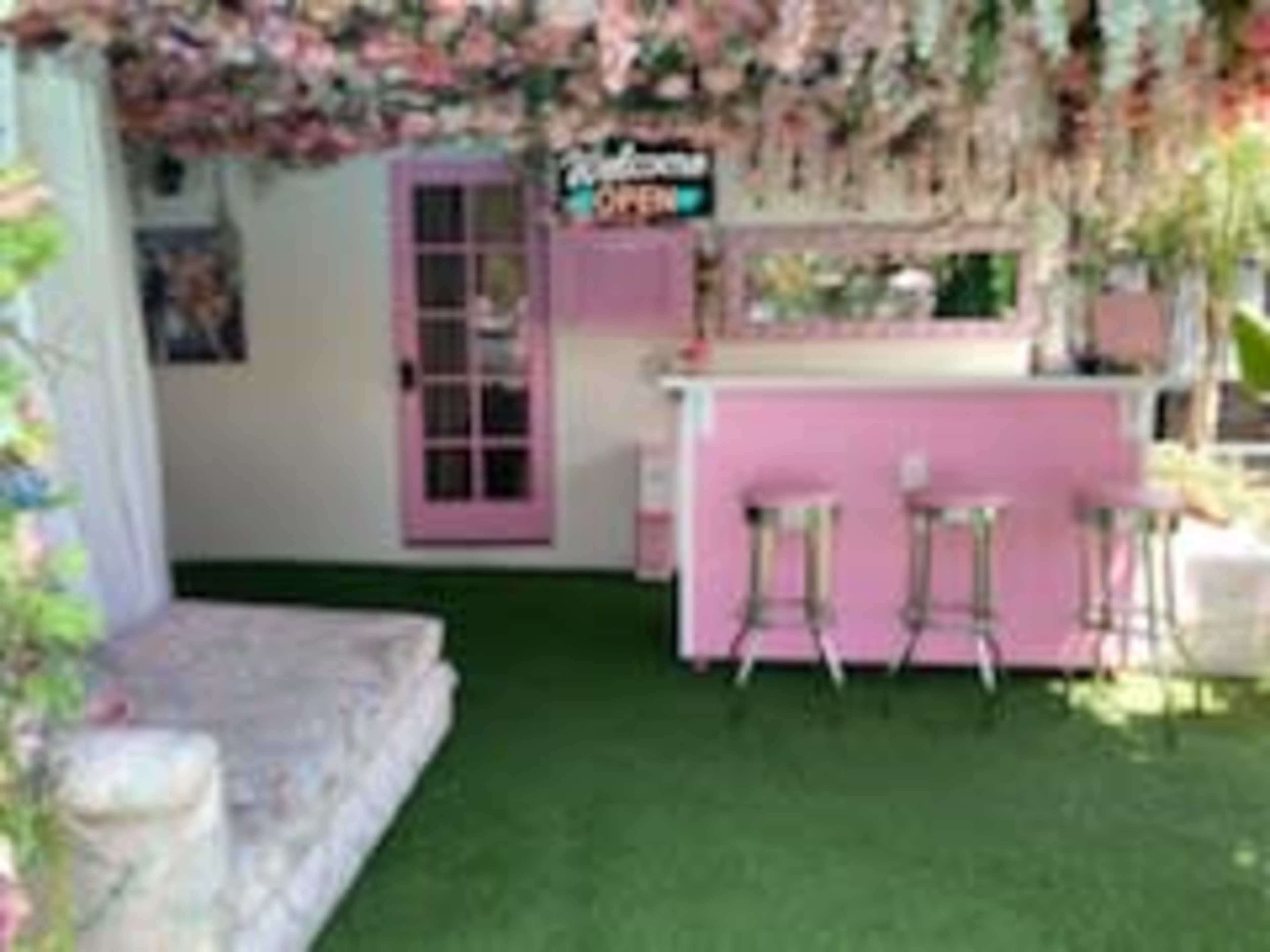 A pink-themed outdoor space with a bar, three stools, and floral decorations overhead.