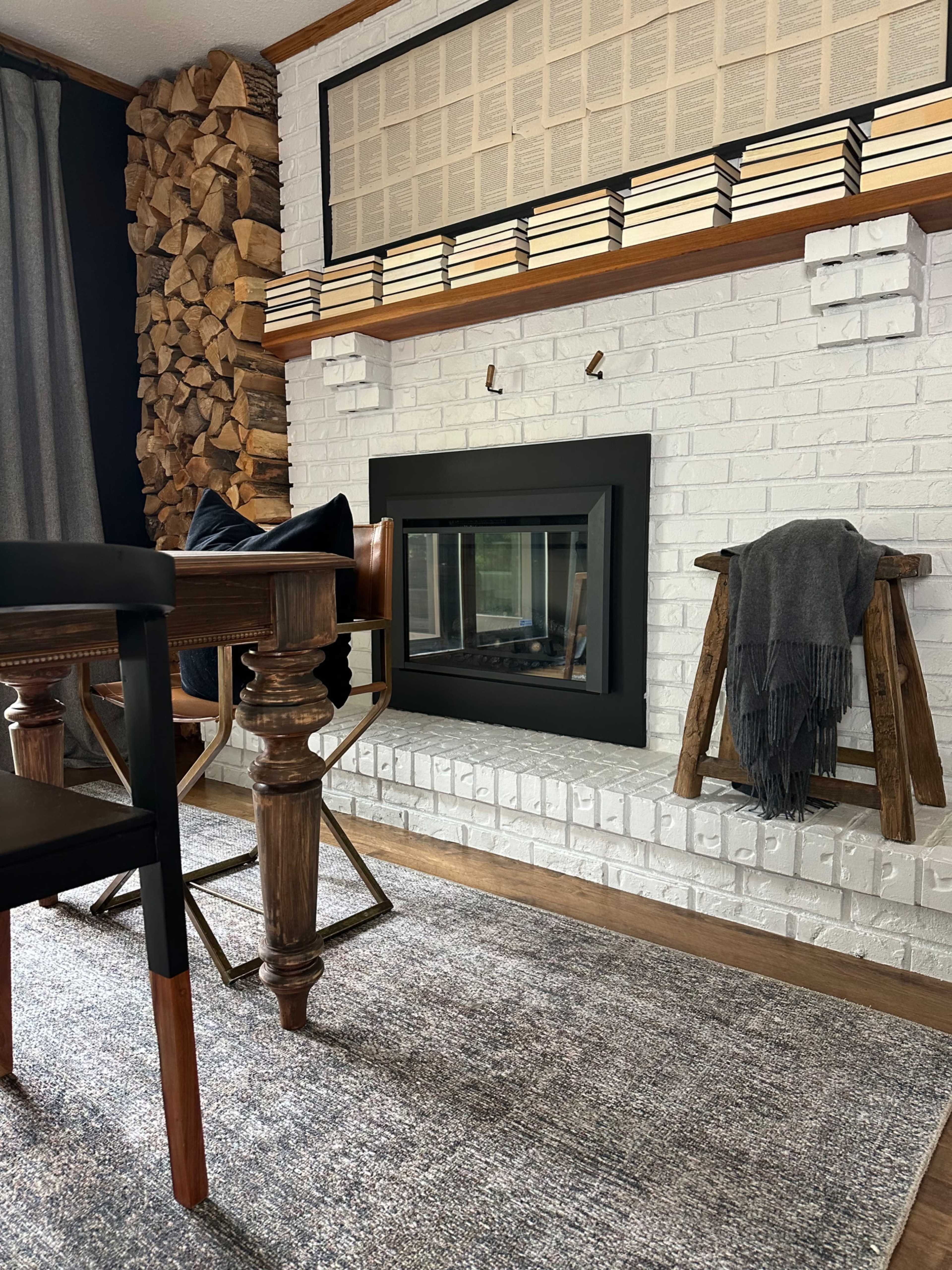 A cozy living room with a brick fireplace, wooden logs stacked beside it, and a dining table with two chairs nearby.