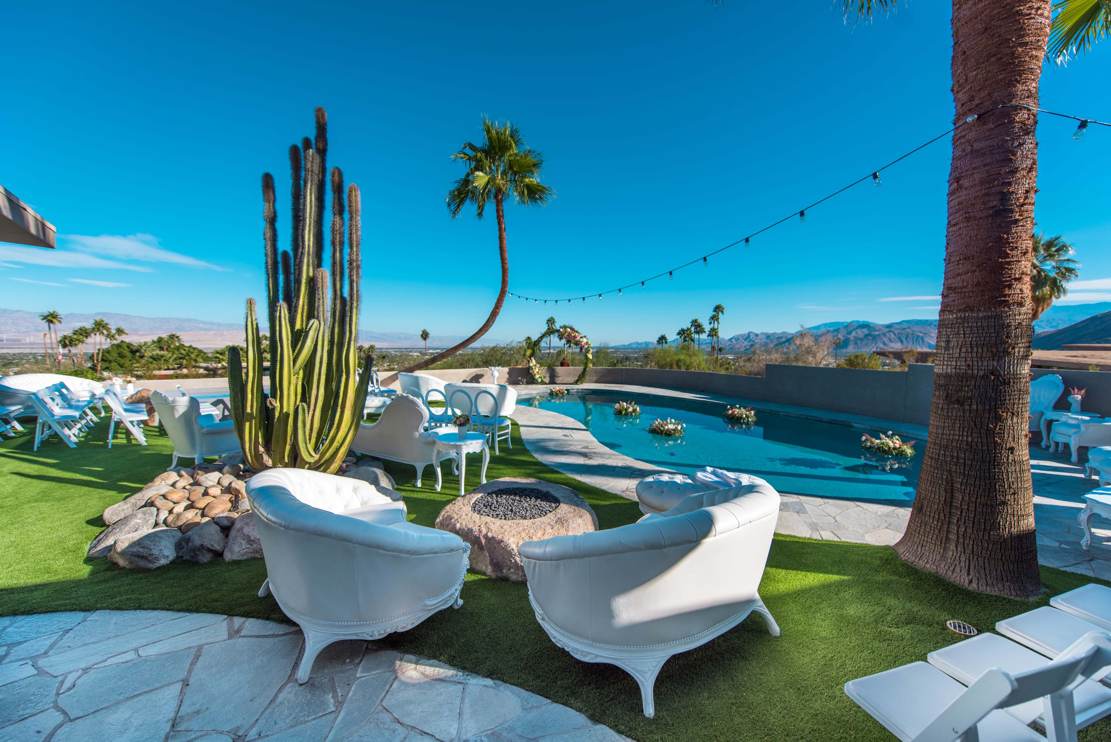 A patio area with white outdoor furniture, a swimming pool, and a large cactus, surrounded by palm trees and mountains in the background.