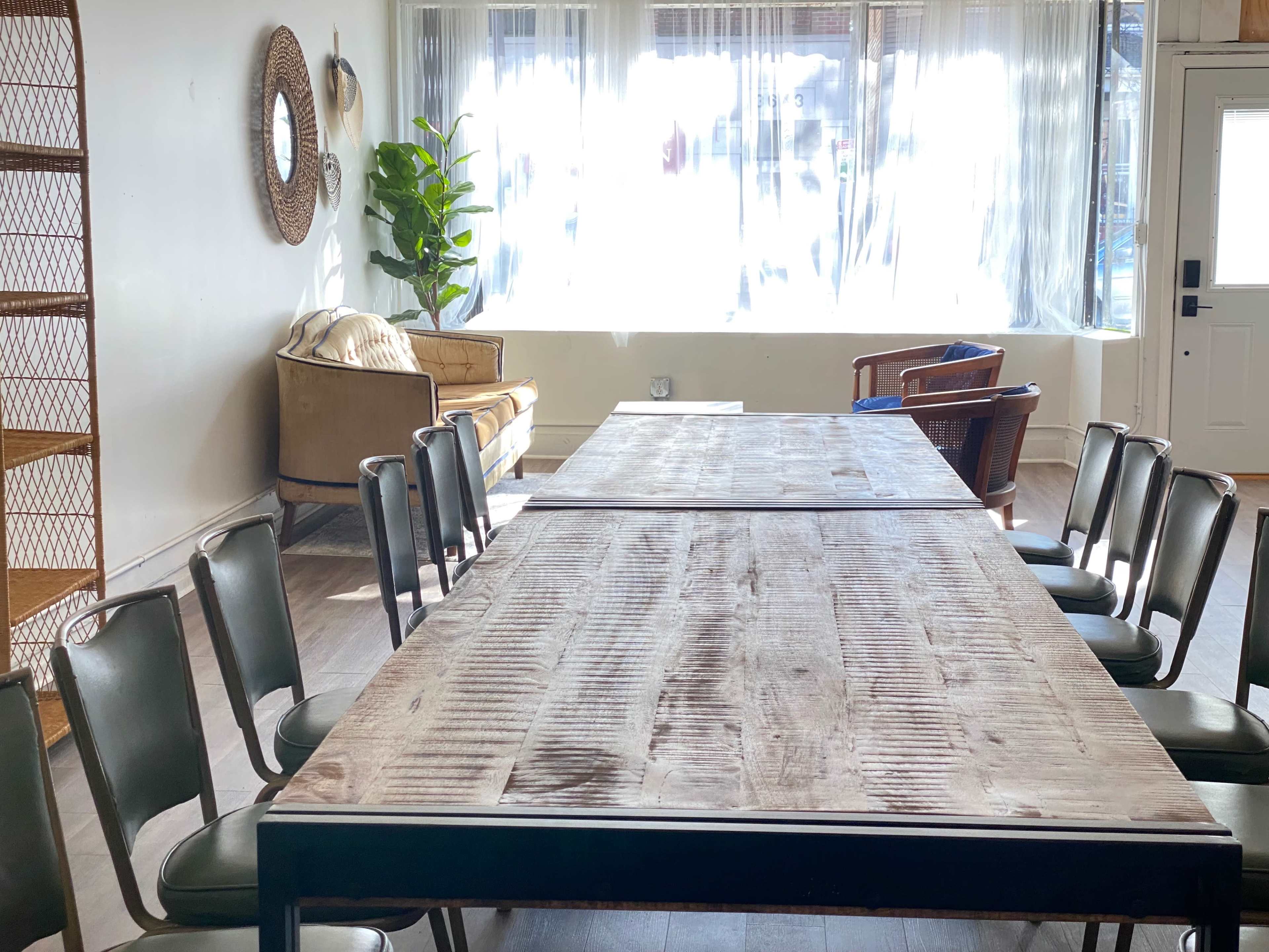A long wooden table is arranged in a bright room with several chairs positioned around it, and a couch in the corner near a window.