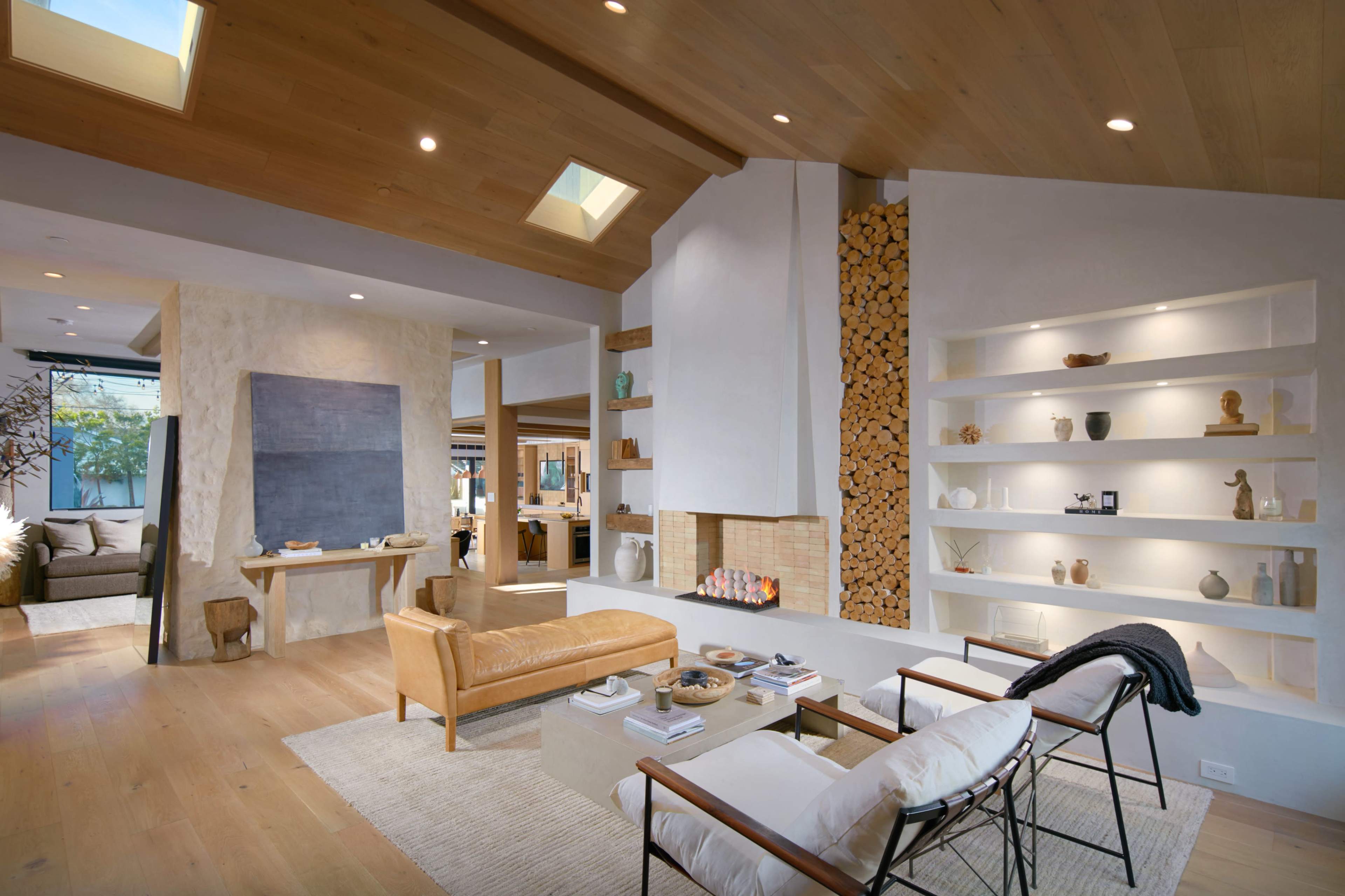 A modern living room featuring a leather sofa, a white armchair, a fireplace with stacked logs, and shelves displaying decorative items.