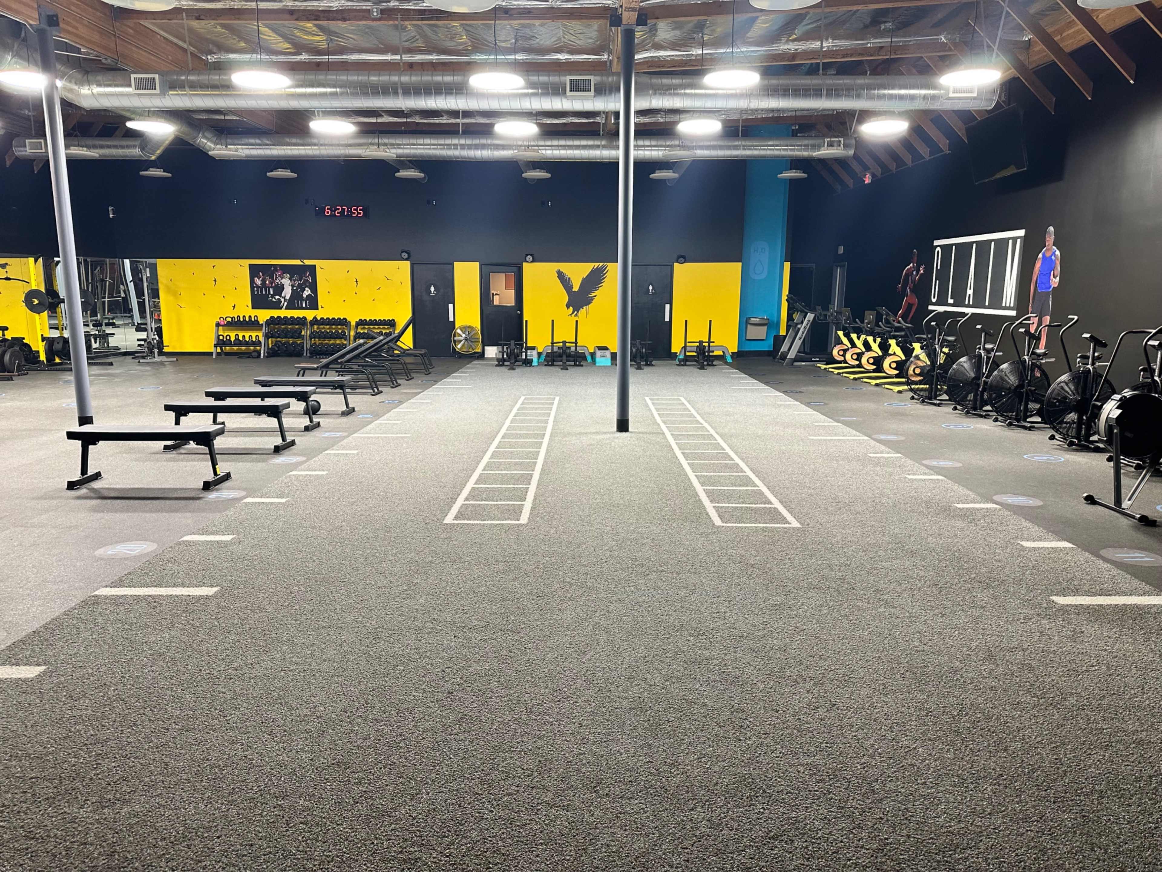 A spacious gym with workout equipment, including benches, weights, and exercise bikes, arranged in a well-lit area with a dark wall and yellow accents.