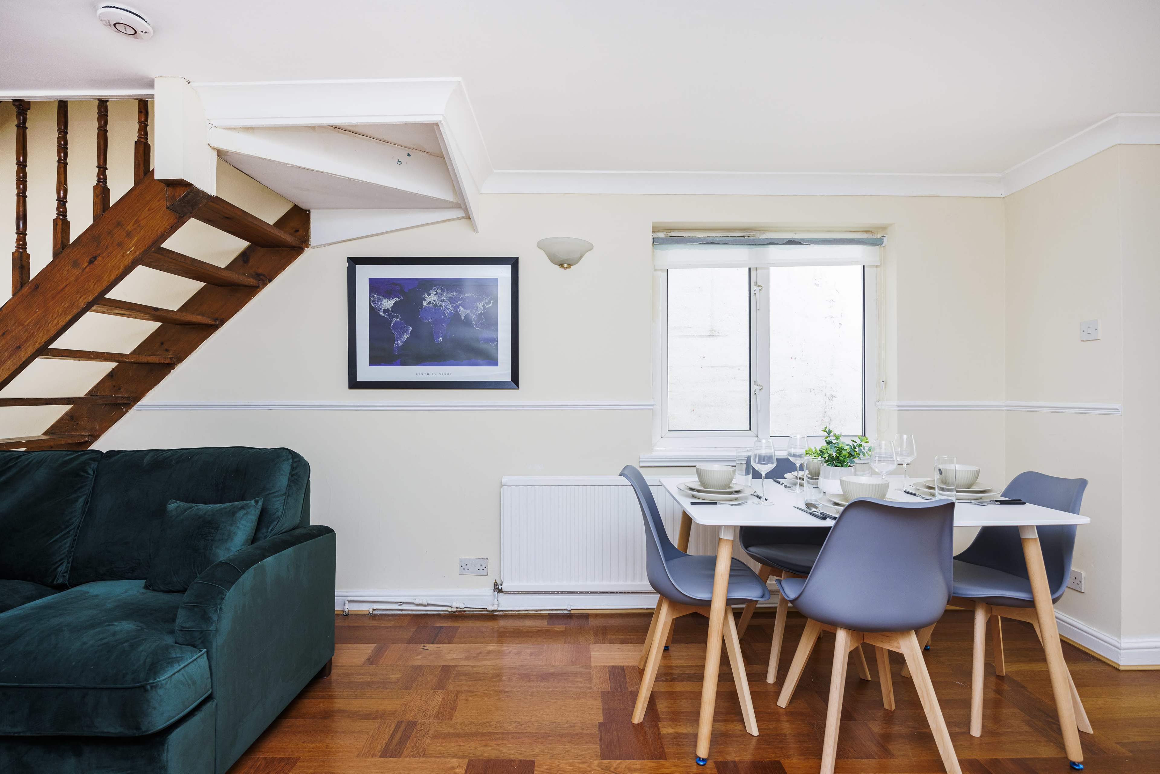 A dining area with a white table and grey chairs is positioned near a window, with a staircase and a green couch visible in the room.