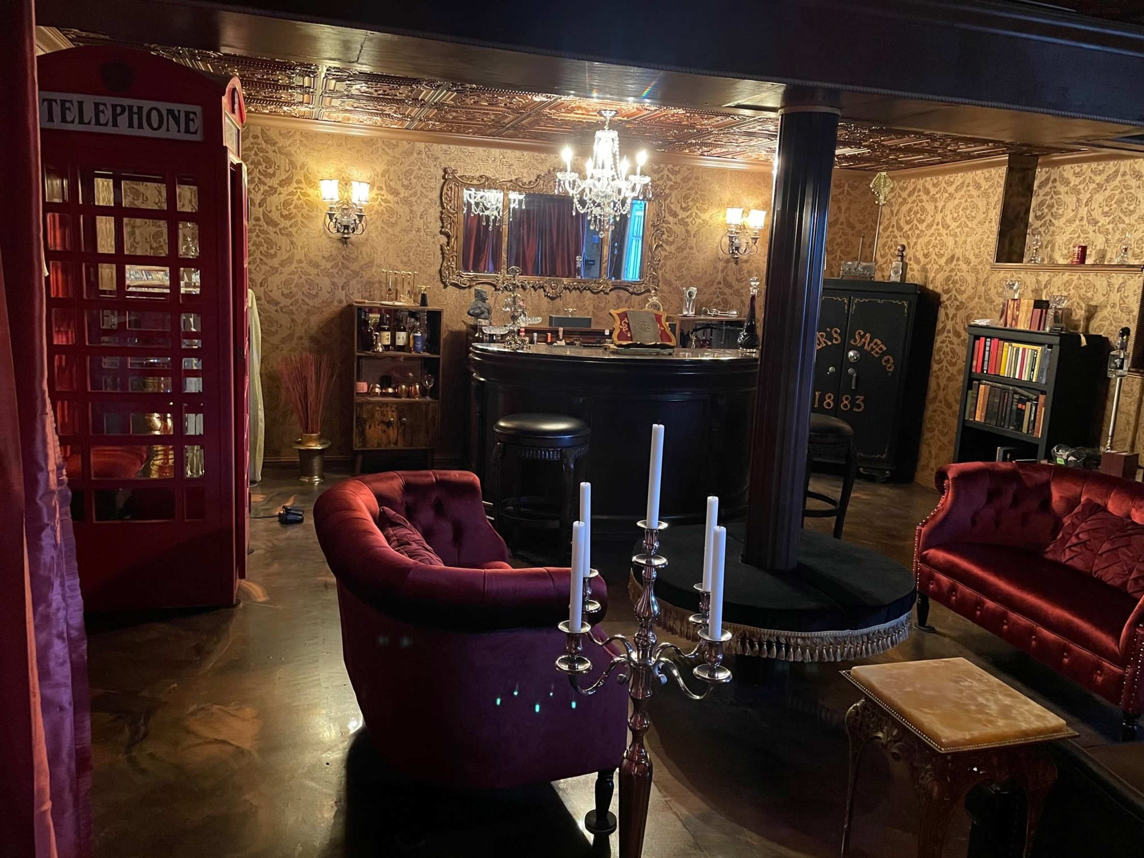 A vintage-inspired room featuring a bar, plush red seating, a chandelier, and a British telephone booth in one corner.