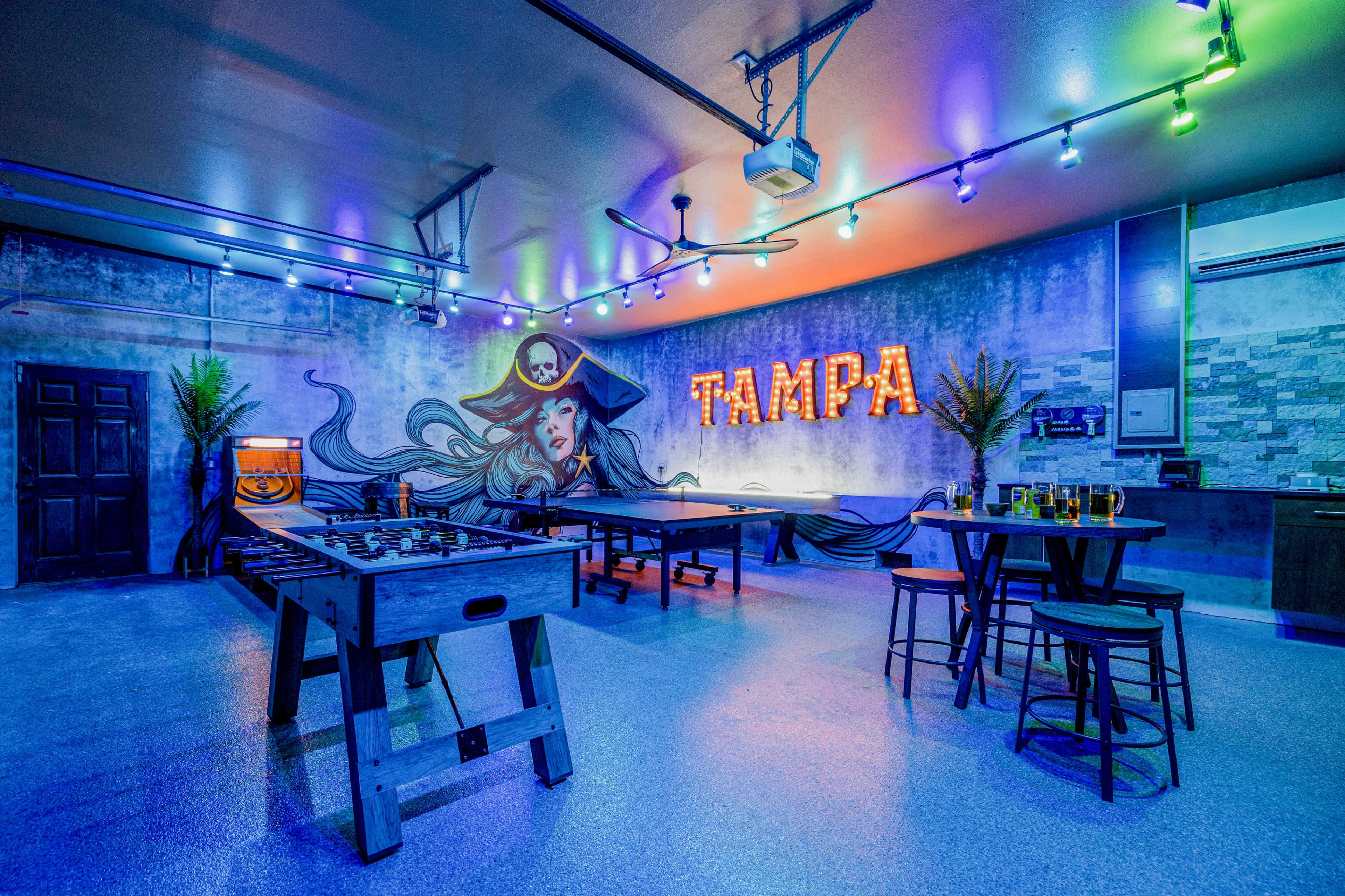 The image shows a vibrant game room with a foosball table, seating area, and a large wall mural featuring a pirate, illuminated by colorful lights and the word "TAMPA" in bright letters.
