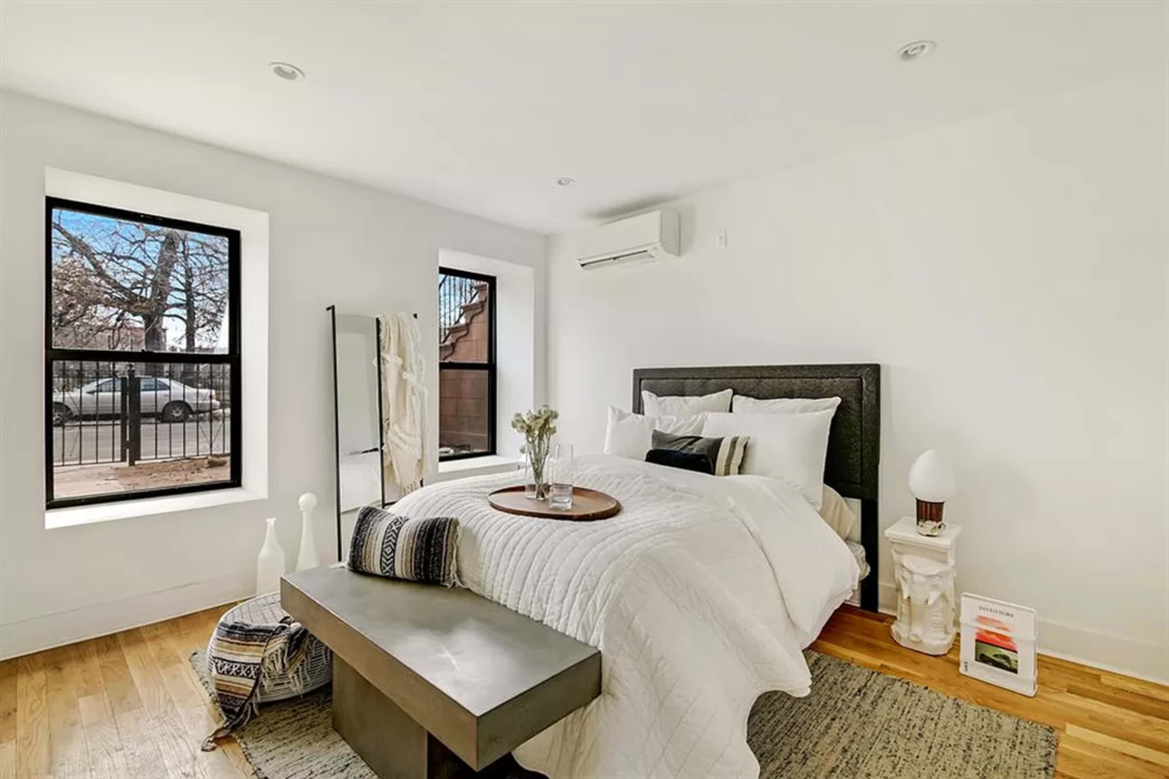 The image shows a minimalist bedroom featuring a large bed with white bedding, two windows with black frames, and a small decorative table next to the bed.