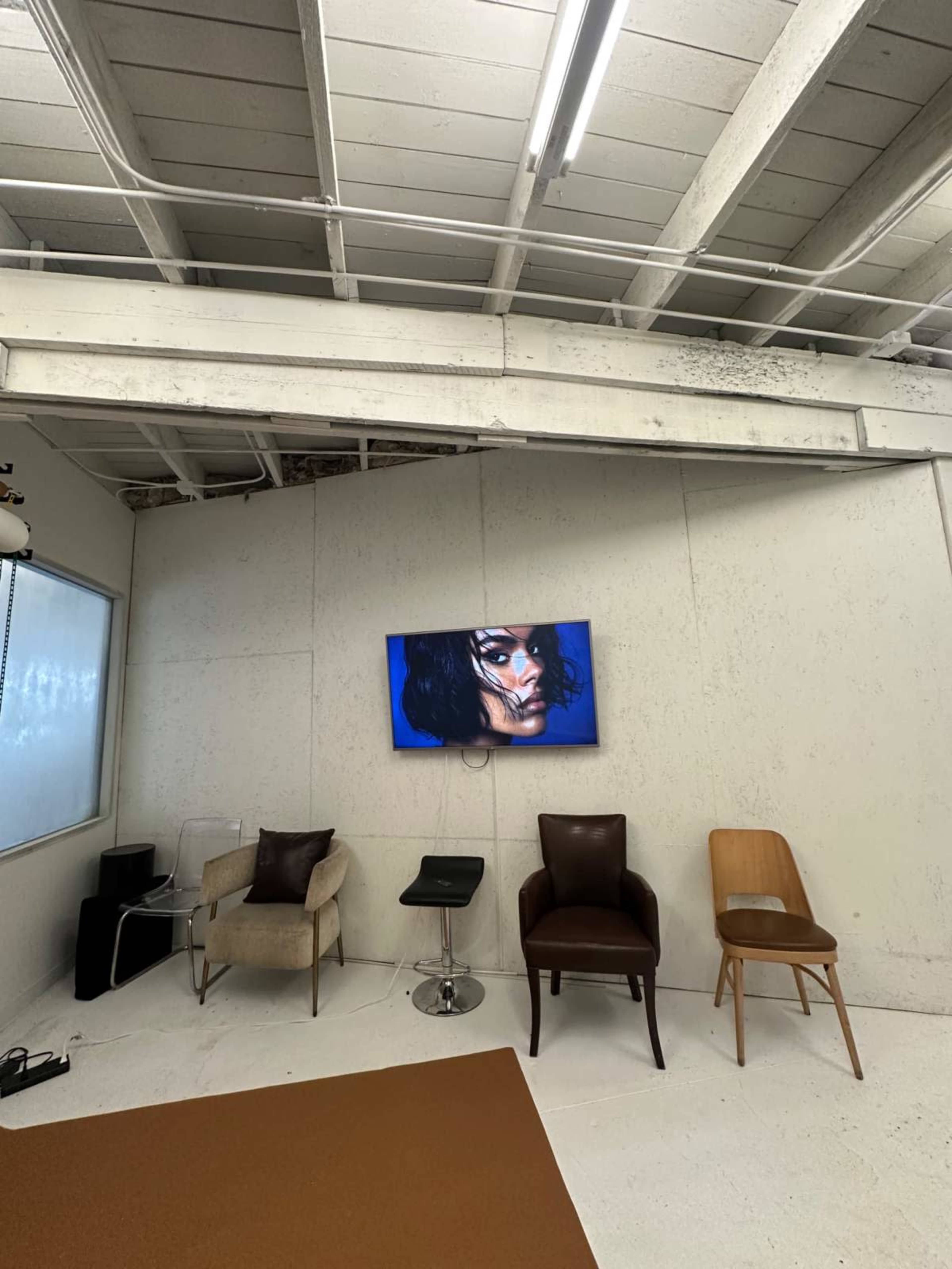 The image shows a minimalist studio space with four chairs and a television mounted on a white wall displaying an artistic image.