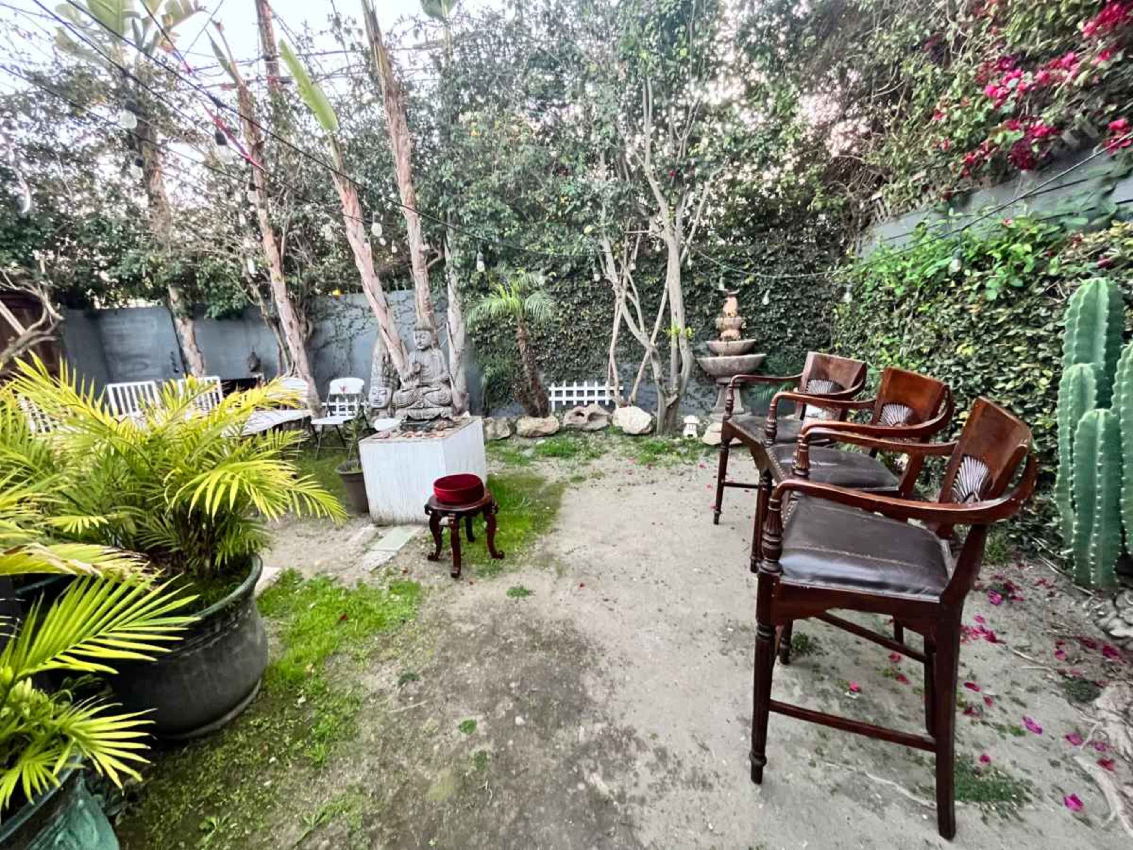 The scene features a serene garden with wooden chairs, potted plants, a small fountain, and surrounded by vibrant greenery.