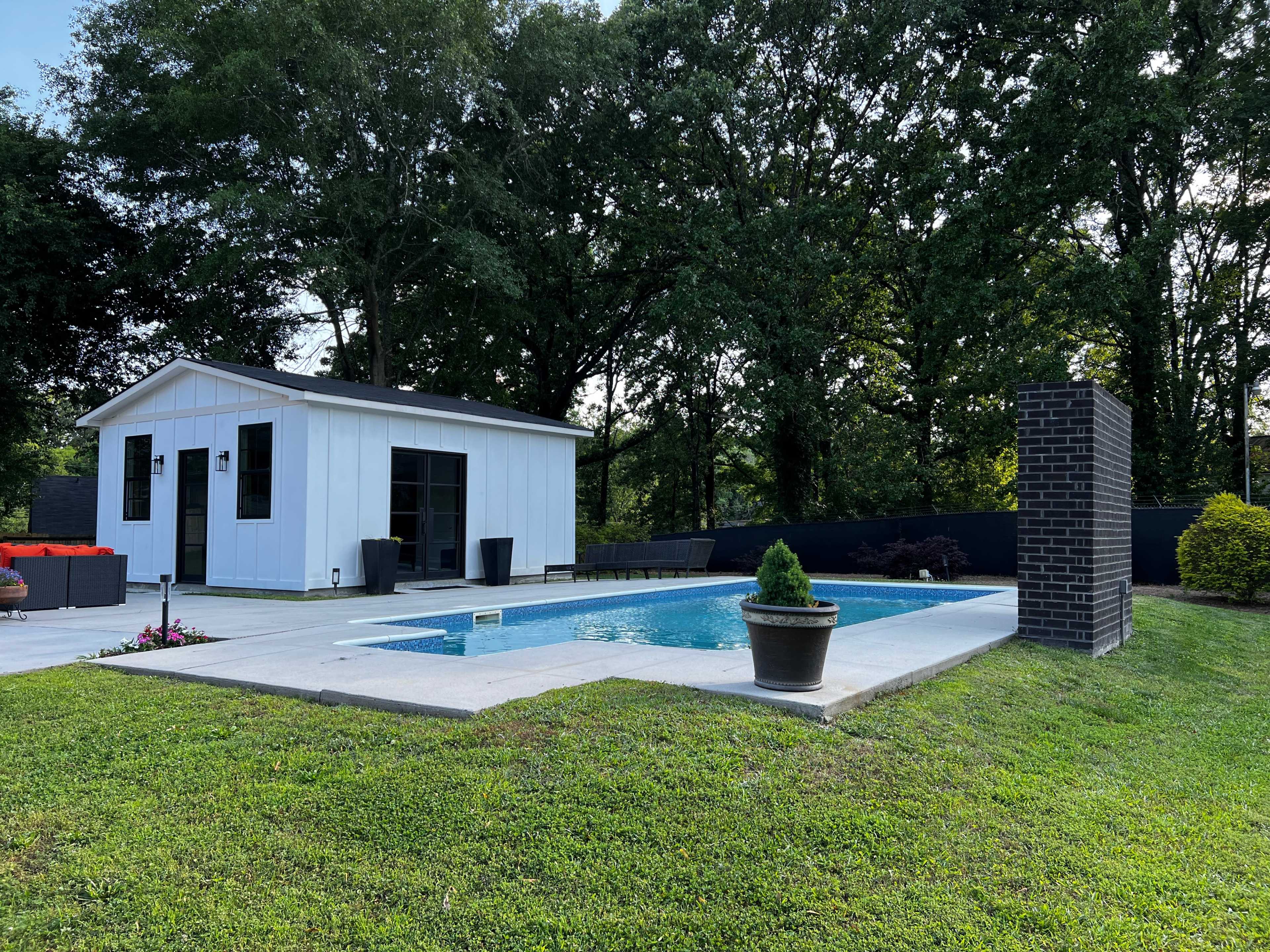 The image shows a modern white shed beside a rectangular swimming pool, surrounded by greenery and potted plants.