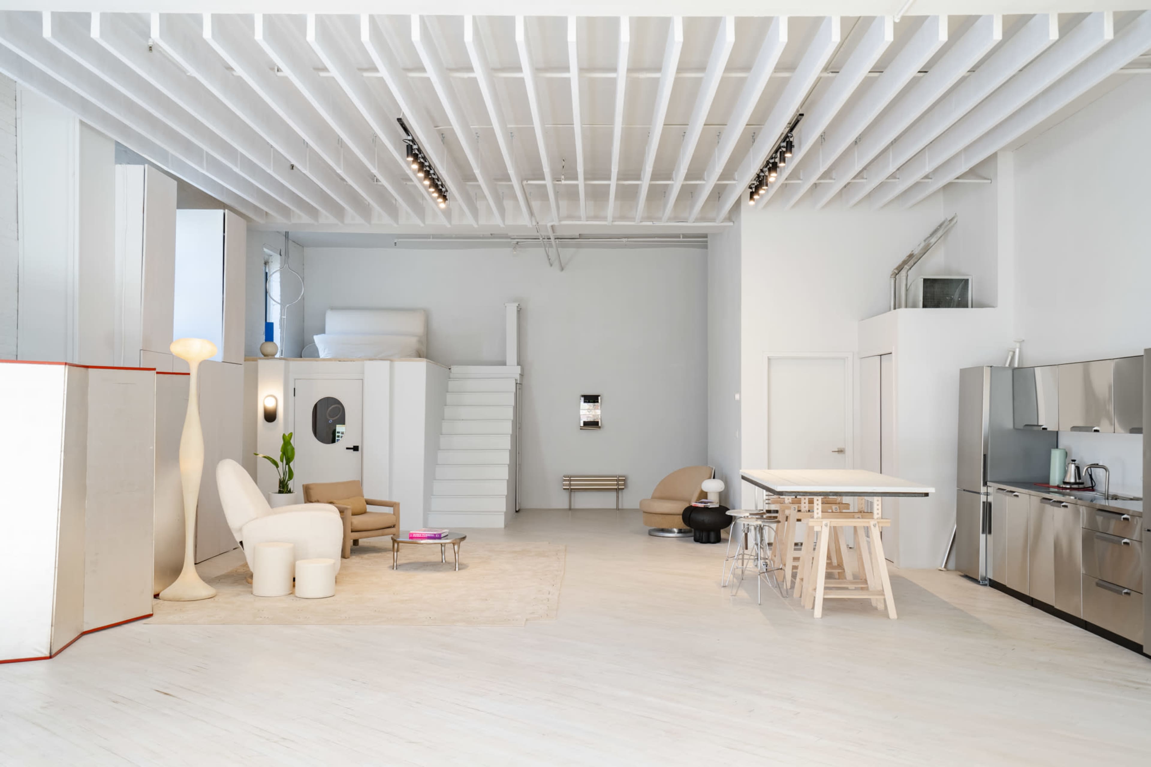 The image shows a spacious, contemporary living area featuring minimal furniture, a kitchen, and a staircase in a well-lit environment.