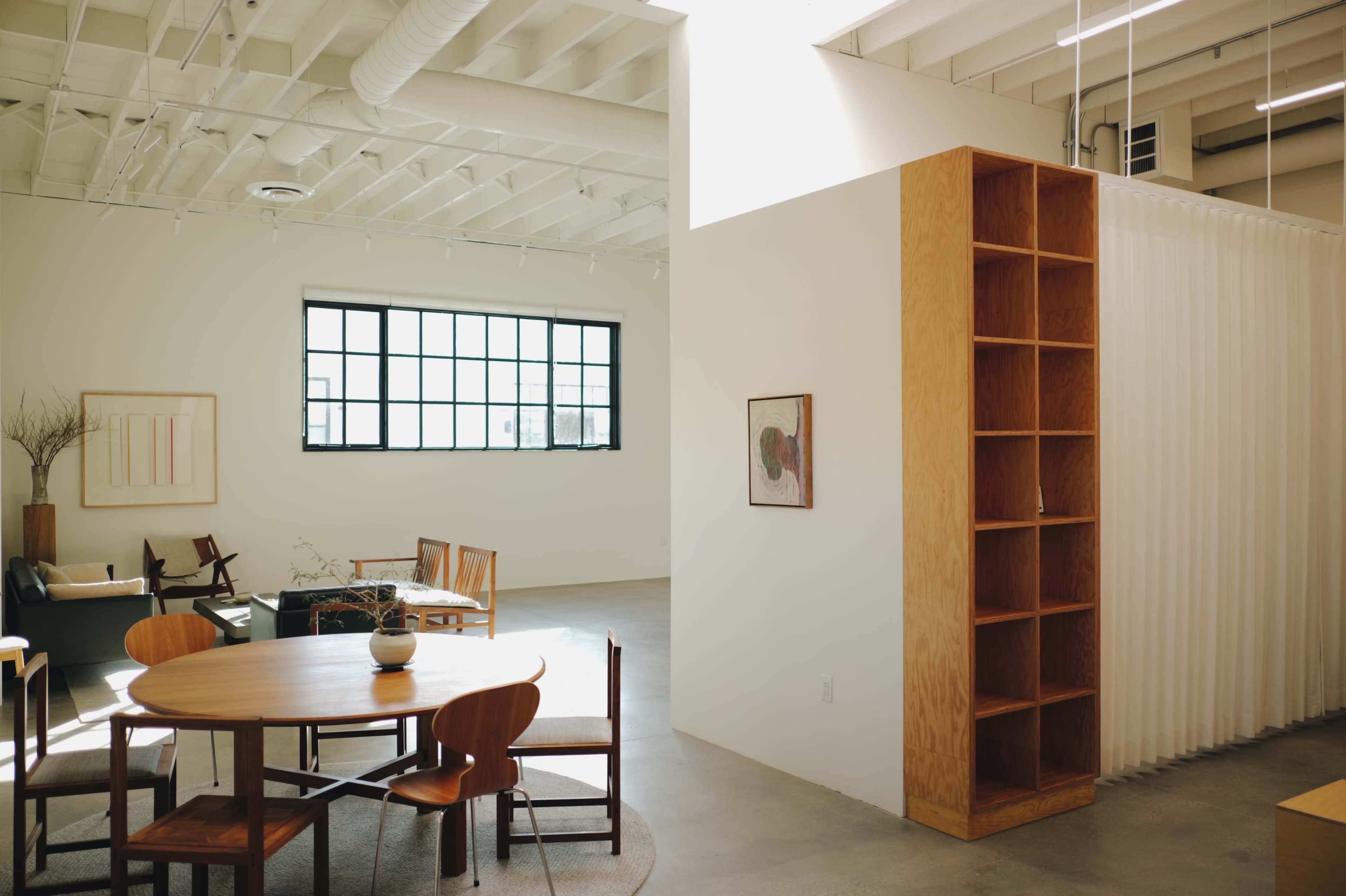 The image shows a spacious, minimalist interior with a wooden dining table and chairs, a tall wooden shelf, and large windows allowing natural light to enter.