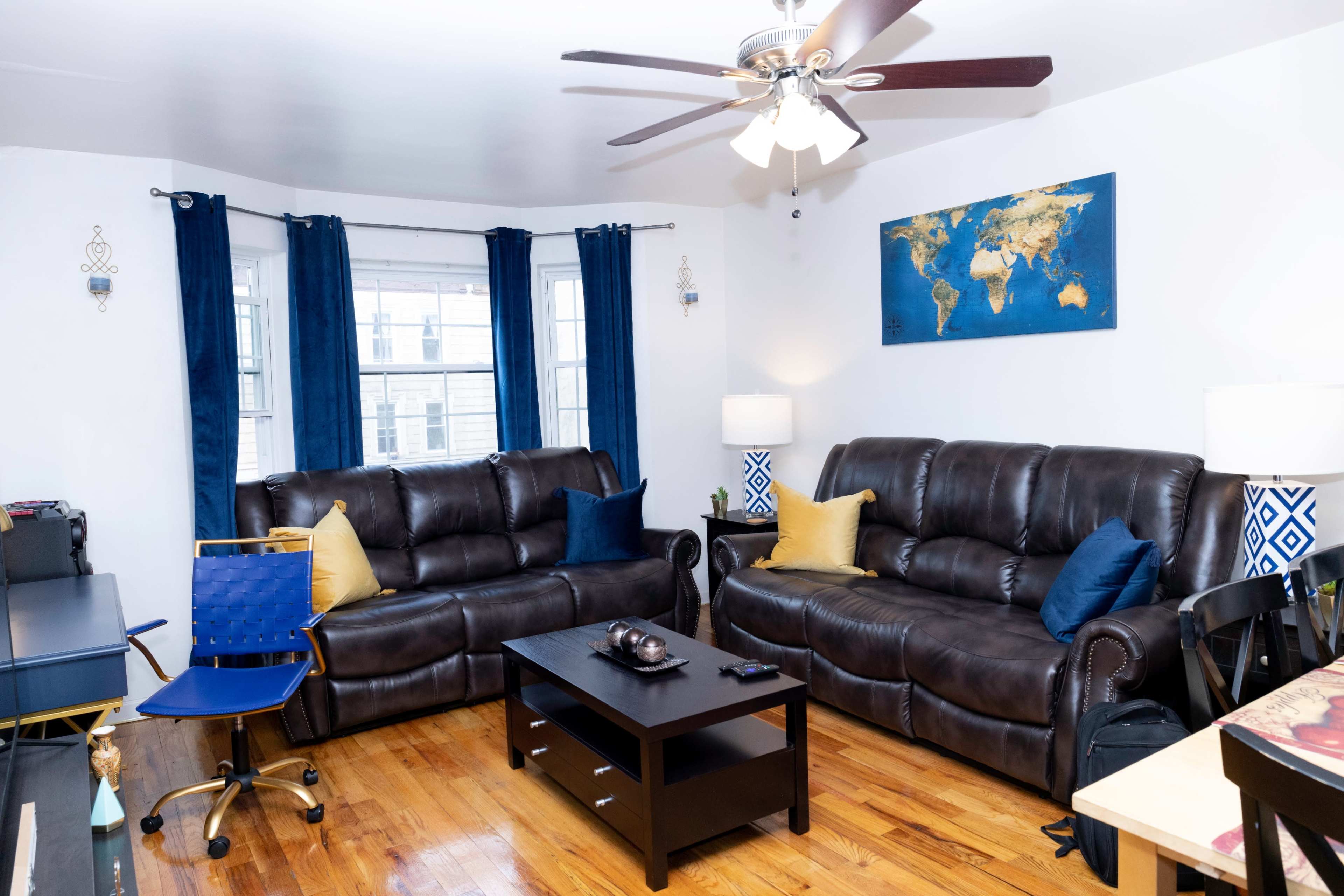 The living room features two brown leather sofas, a black coffee table, a blue office chair, and a world map on the wall, with large windows dressed in blue curtains.