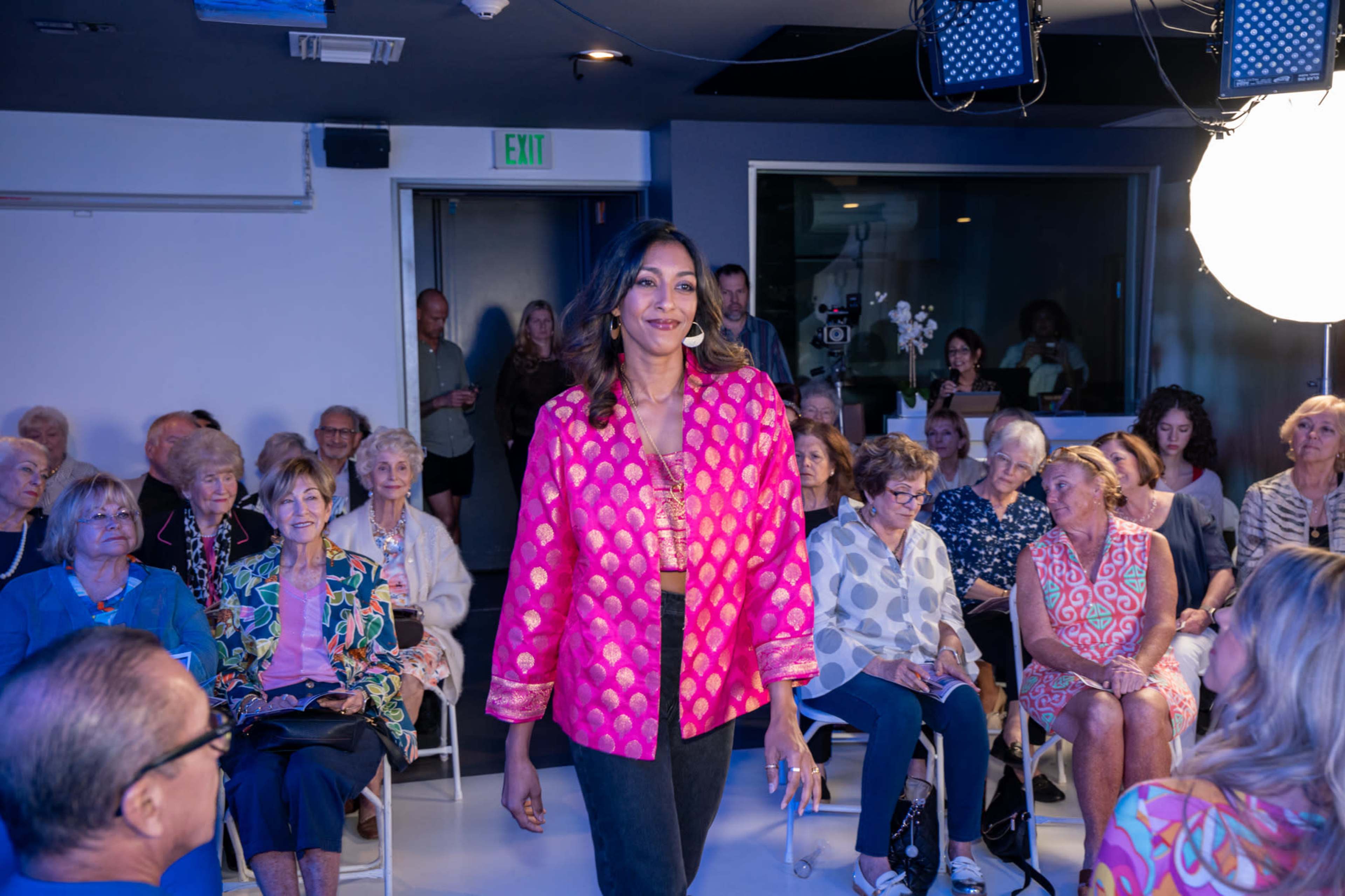 A model walks down a runway in front of an audience seated in a brightly lit room.